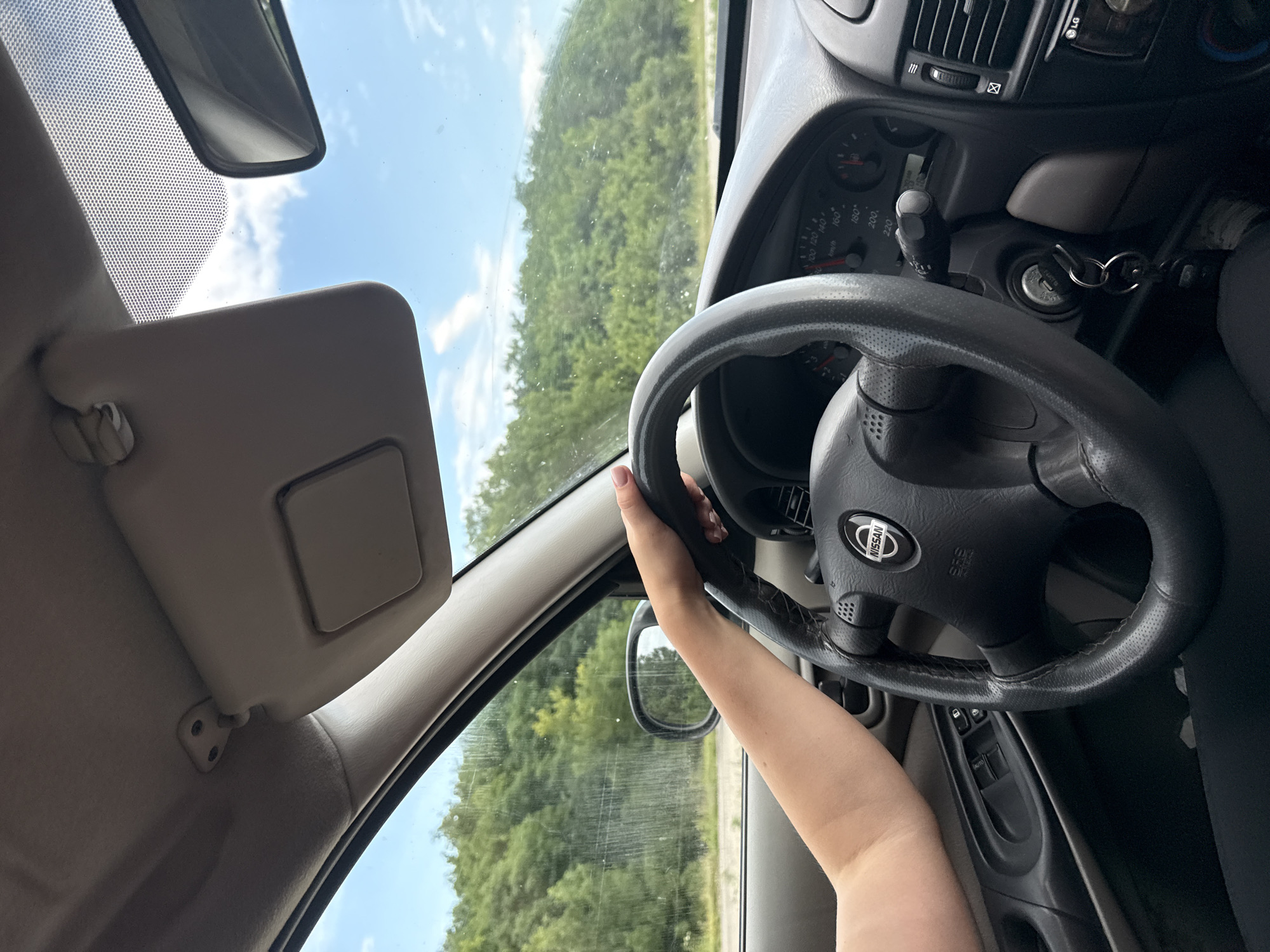 An interior view from the passenger seat of a moving car shows a person's left arm and hand on the steering wheel. The steering wheel is black leather with a Nissan logo in the center. The dashboard has a speedometer and other gauges. Outside the car, a green, wooded hillside is visible under a cloudy blue sky. The scene appears to be on a road outside of Razeni, Moldova, based on the metadata.