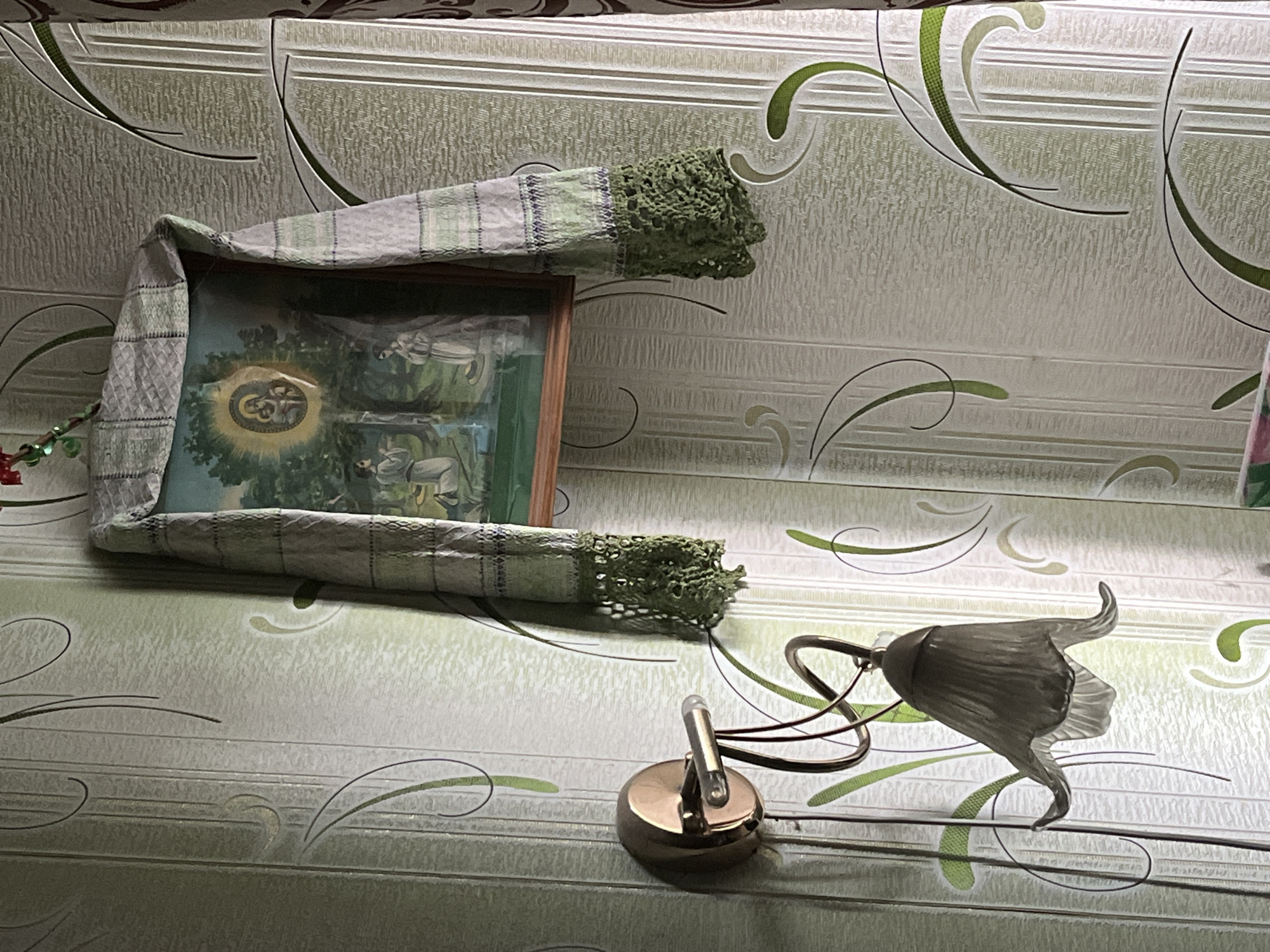 The image displays a section of an indoor wall adorned with light-colored wallpaper featuring a pattern of horizontal lines and decorative green and black swirling motifs. Centered on the wall is a framed picture, appearing to be a religious icon or painting, partially covered by a light-colored fabric. This fabric, possibly a checkered or striped towel with a green crocheted trim, is draped over the top and sides of the wooden frame. The visible part of the icon depicts figures in a lush, green landscape, with a prominent circular, glowing emblem at its center. Within this emblem, intertwined Cyrillic letters are visible, appearing to spell "БОГЪ" (God).
To the left of the framed picture, small red and green decorative elements can be seen, possibly part of a garland. Below and to the right of the icon, a two-part wall-mounted light fixture is present. It consists of a round, brass-toned base supporting a small, clear, cylindrical bulb. A separate, darker metallic arm extends from the wall, holding a larger, frosted or textured glass lampshade shaped like an inverted flower or bell.
No people are visible, and no specific activity is taking place beyond the static display of these decorative and religious items. The setting is an interior space, consistent with a home in Minsk, Belarus, where icons are often displayed and adorned in this manner. The time of day and weather are not discernible from the image.