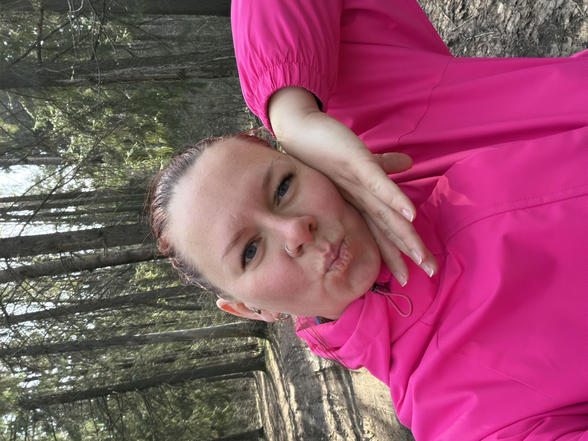 A woman is taking a selfie outdoors. She has red hair pulled back from her face, fair skin, blue eyes, and a nose ring. She is wearing a bright pink jacket. Her left hand is near her face, with her fingers slightly curved. The background consists of a forest with numerous trees. The location is Chaikovsky, Russia.