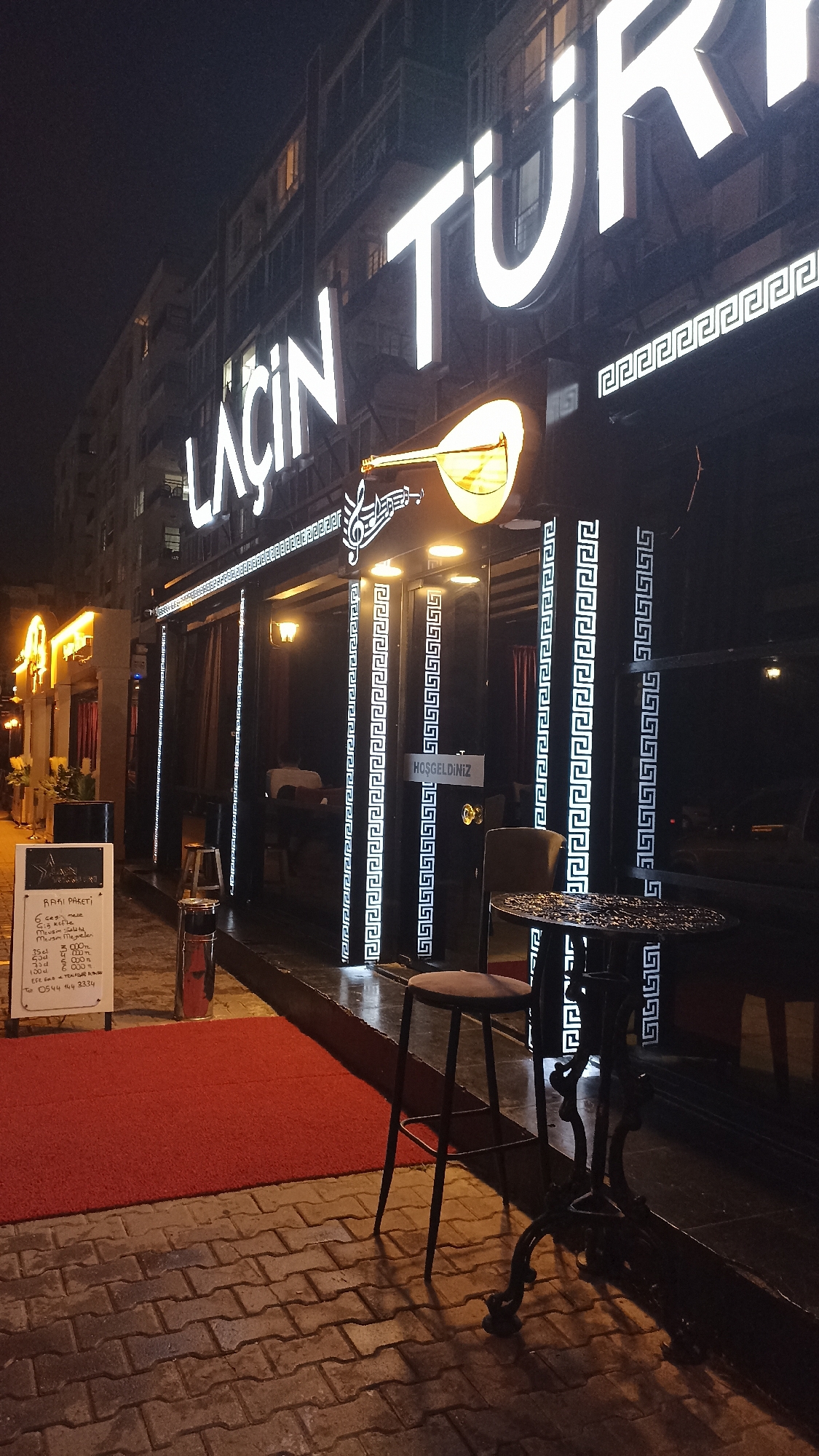 The image captures the illuminated exterior of a building at night, likely a restaurant or bar, in an urban setting in Yenişehir, Turkey. The prominent sign on the facade reads "LAÇIN TÜR" in large, bright white letters, accompanied by an illuminated emblem of a saz or baglama (a traditional Turkish string instrument) and musical notes. Decorative patterns resembling the Greek key (meander) adorn the building's front, framing dark glass panels that serve as windows or doors.
A small, ornate black metal table and a matching stool are placed on the sidewalk in front of the entrance. A red carpet leads from the pavement towards the establishment. A white sandwich board-style menu, written in Turkish, is visible on the left. The text on the board includes "RAKI PAKETI" (Raki Package), detailing offers such as "6 çeşit meze" (6 kinds of appetizers), "Mevsim Meyveleri" (Seasonal Fruits), and various Raki bottle sizes (35cl, 50cl, 70cl, 100cl) with corresponding prices in Turkish Lira (TL): 300 TL, 400 TL, 500 TL, and 600 TL. It also mentions "Efe Rakı" and a phone number: "0544 144 3334". The door features a sign that reads "HOŞGELDİNİZ" (Welcome).
The scene is lit by the establishment's signage and warm-toned lights, creating an inviting atmosphere typical of an evening venue. The street is paved with interlocking bricks. No people are directly visible in the image. The time of day is night, indicated by the dark sky and artificial lighting. The overall impression is that of a cultural dining and entertainment spot.