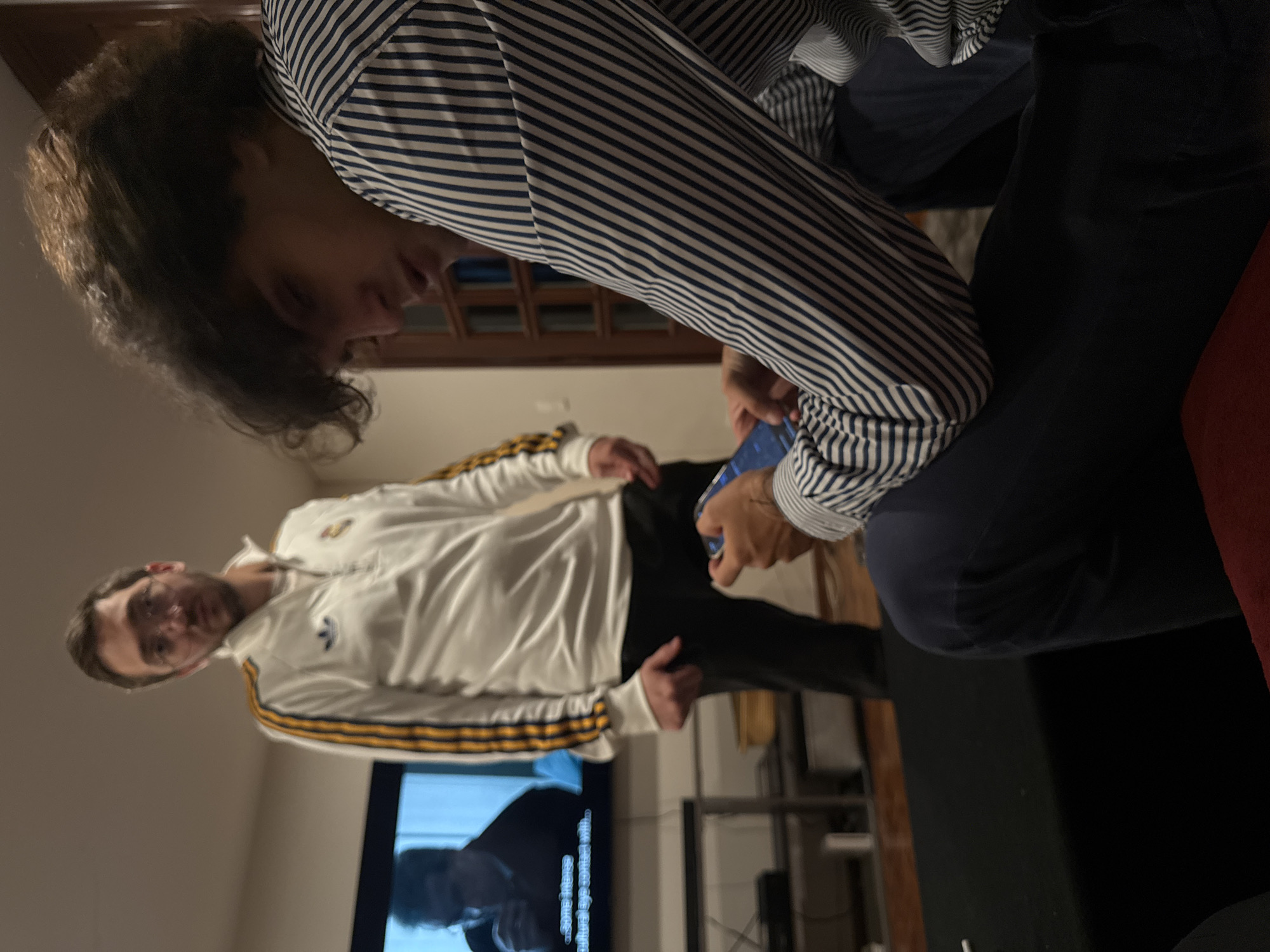 Two men are indoors. One is wearing a striped blue and white shirt and dark pants, looking down at a blue object in his hands. The other man is wearing a white tracksuit with yellow stripes and is standing near a television. The television shows a blurred image. The location is indoors, likely a home. The city is not discernable from the image.