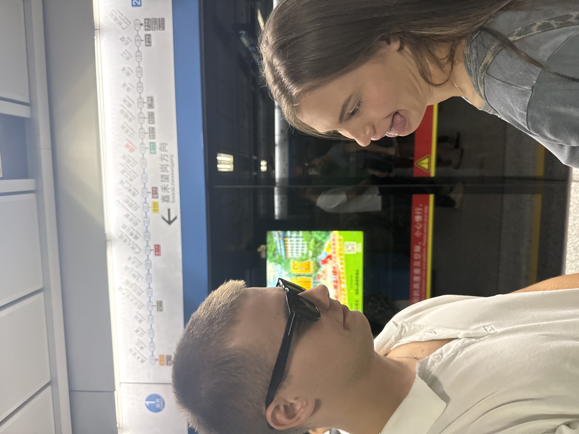 In Haizhu District, China, a Caucasian man wearing sunglasses and a white polo shirt stands to the left, facing a Caucasian woman who is sticking her tongue out at him, positioned to his right. Both individuals appear to be on a subway platform, evidenced by the presence of subway line maps and train doors behind them. The map displays various station names, including what appears to be "嘉禾望岗方向" (Jiahewanggang Direction). The background also shows a screen with colorful images and text in Chinese.