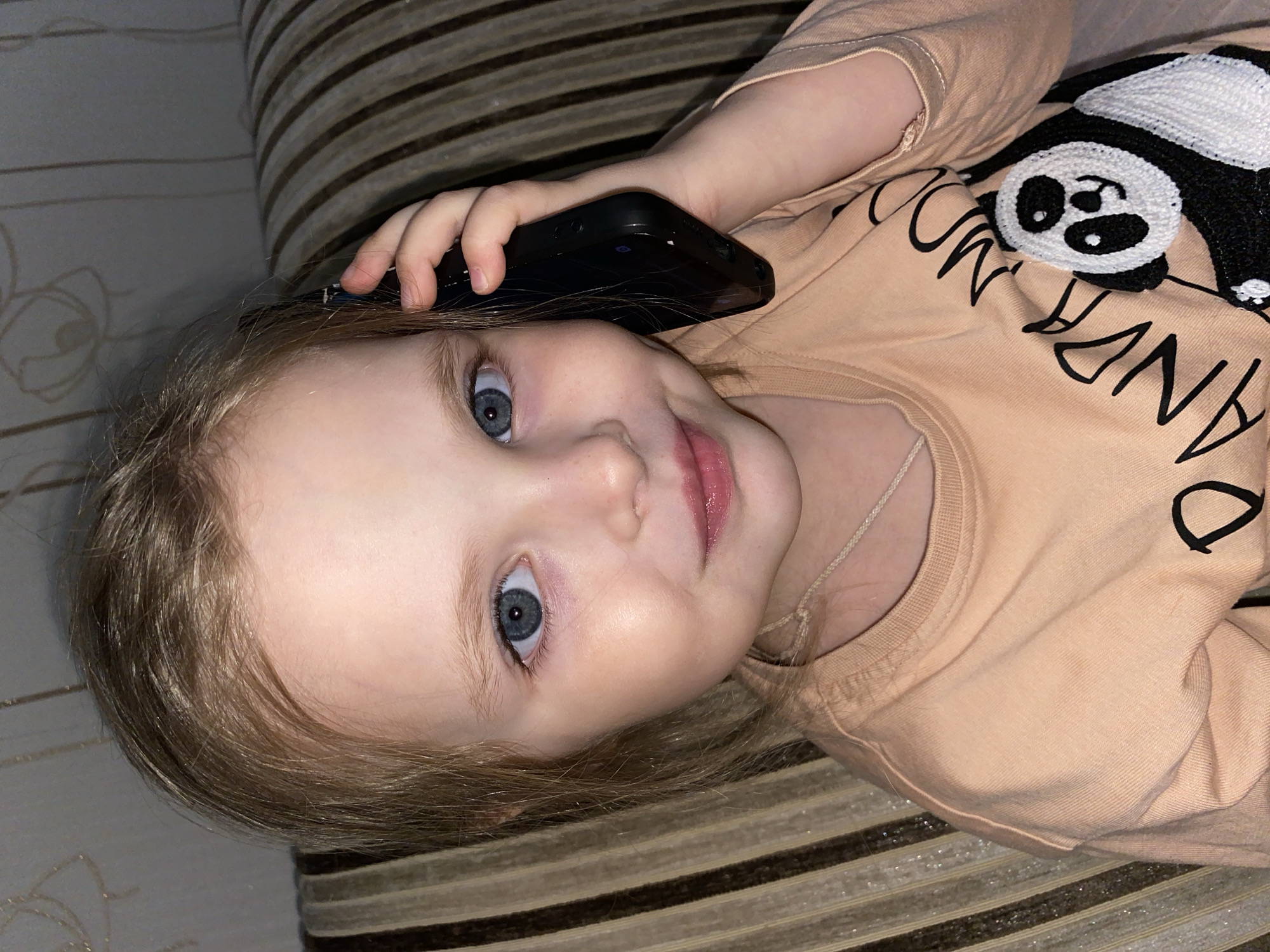 The image captures a young child, likely a girl, lying on her back indoors, holding a black smartphone to her right ear. She has fair skin, light blonde hair, and wide, blue eyes that are looking directly at the camera with a soft, curious expression and a slight smile. Her head is positioned towards the left side of the frame.
She is wearing a peach-colored, long-sleeved shirt. The shirt features a prominent black and white knitted panda graphic on the upper right side. Below and to the left of the panda, black text is visible, clearly showing "PANDA" and partially visible letters that appear to spell "MOOD," suggesting the phrase "PANDA MOOD" or similar.
The background behind the child consists of two distinct patterns. Immediately behind her head and body is a dark and light striped fabric, likely the upholstery of a sofa or bed. Further back and to the left, a lighter-toned wall with a subtle, possibly floral or damask, pattern can be seen. The overall lighting is consistent with an indoor setting, possibly with artificial light. The activity depicted is the child playfully or genuinely engaging with the phone, mimicking a call. The image does not contain any visual cues about the time of day or weather. While the context suggests the location is Votkinsk, Russia, this detail is not discernible from the visual content of the image itself.