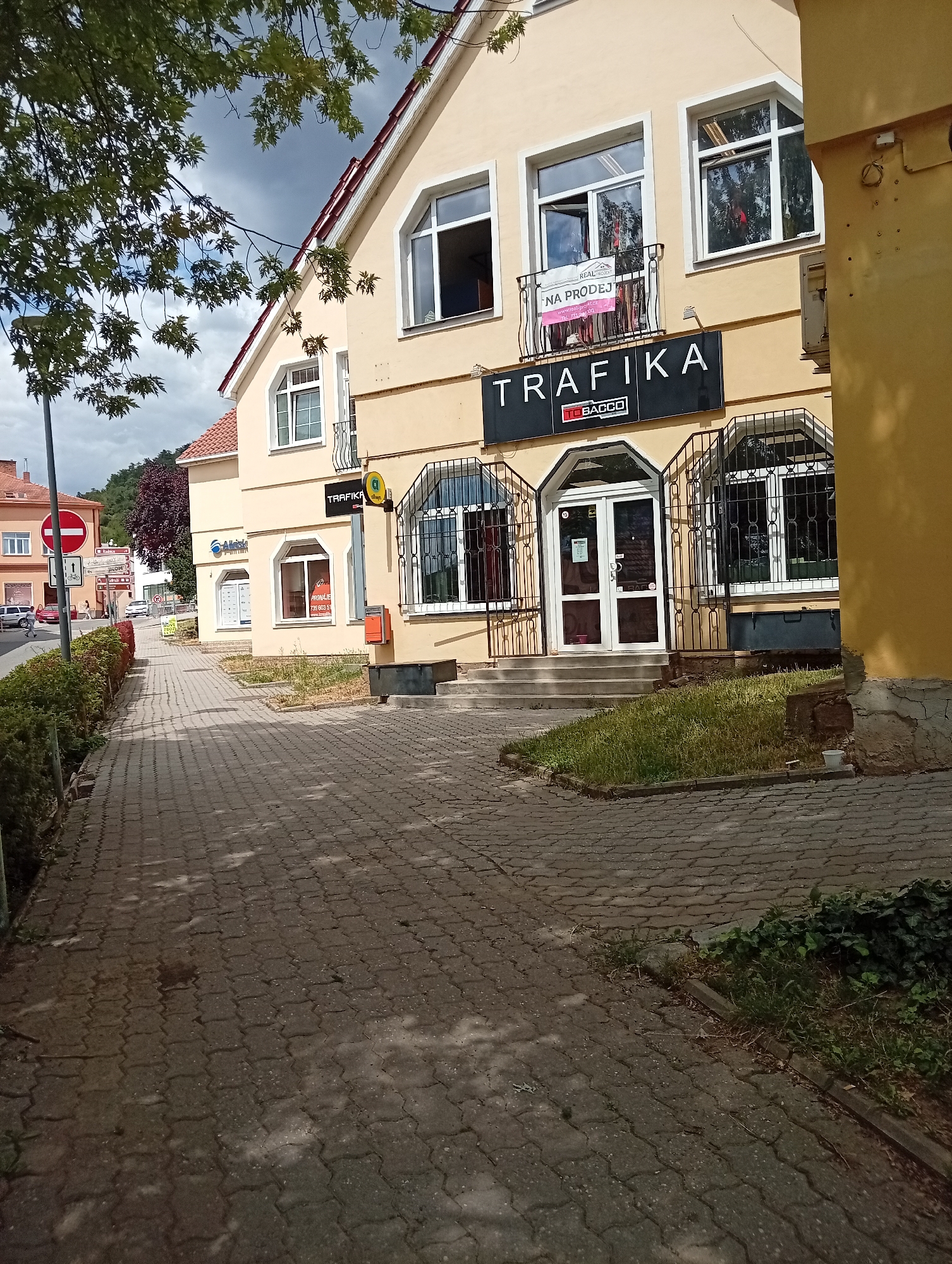The image shows a street scene in Tišnov, Czechia, on a cloudy day. A light yellow building with a red roof dominates the foreground, featuring a store called "TRAFIKA". Above the store entrance, a balcony displays a pink sign that reads "NA PRODEJ" (For Sale). The building has several windows, some with decorative metal grills. A paved sidewalk runs along the building, bordered by hedges and grass. Across the street, other buildings and a traffic sign are visible. A few cars are parked along the street. The overall impression is of a quiet, somewhat aged, commercial area.