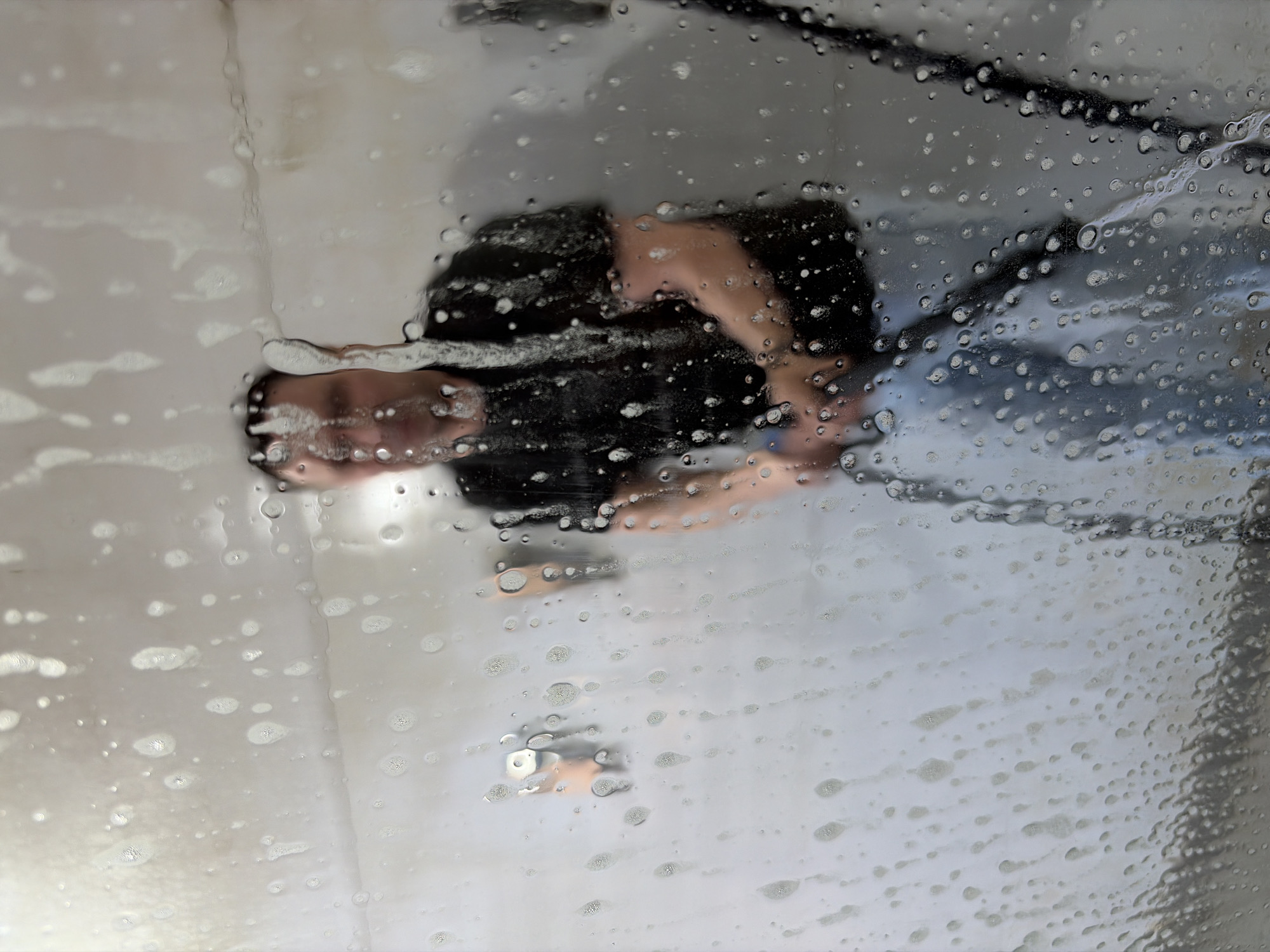 A slightly blurred, high-angle shot presents a view through a wet window or glass surface, partially obscured by numerous water droplets and streaks of soap residue. The reflection of a person is visible through the wet glass. The individual appears to be wearing a dark top and a light-colored lower garment, possibly jeans. The face of the person is visible, although distorted by the water droplets, making precise facial details indistinct. The person seems to be looking down, possibly at the surface being cleaned. A black cable or hose is also visible near the top of the frame. The background is blurry and undifferentiated, with no indicators of the specific location in Izhevsk, Russia. There is no other contextual information discernible from the image.