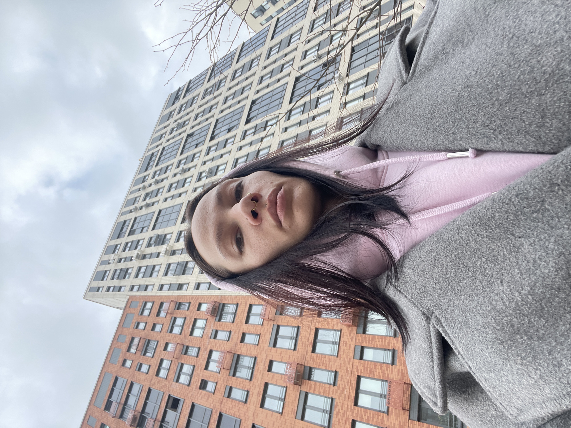 A low-angle, tilted selfie features a young adult with dark hair, a nose ring, a pink hoodie, and a gray coat. Behind the person are two multi-story buildings. The taller building has a gray facade with many windows. The shorter building is red brick with similar windows. The cloudy sky is visible in the upper left corner. The buildings suggest an urban setting. While the city and exact location are not identifiable, the user has specified it is Izhevsk, Russia.