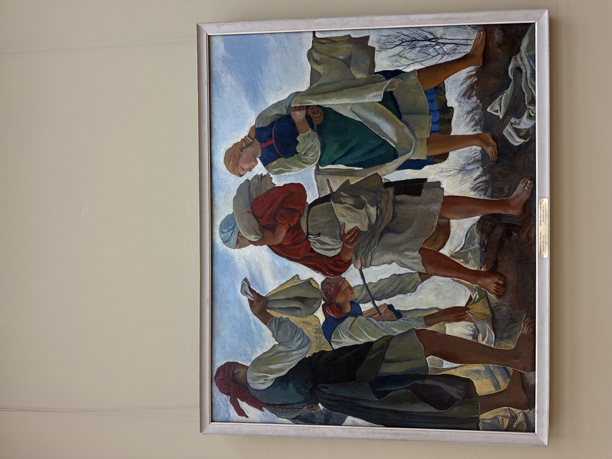 The image is a painting in a silver frame hanging on a beige wall. The painting depicts four figures walking in a row, facing in the same direction. The figures appear to be peasants or working-class individuals, based on their clothing. The topmost figure wears a dark blue top, a green dress, and a white shawl. The second figure wears a red top and a greyish shawl. The third figure wears a white shirt and a light blue piece of clothing over her head, while the bottom figure wears a greyish white shirt, a blue top, and a black skirt. All figures are barefoot. The background includes a blue sky, what appears to be leafless trees, and a rocky or muddy ground. There is a label in Russian on the right side of the frame. The painting is identified as "SEREBRIAKOVA Z. YE. 1884-1967" which is in the State Tretyakov Gallery in Moscow, Russia.