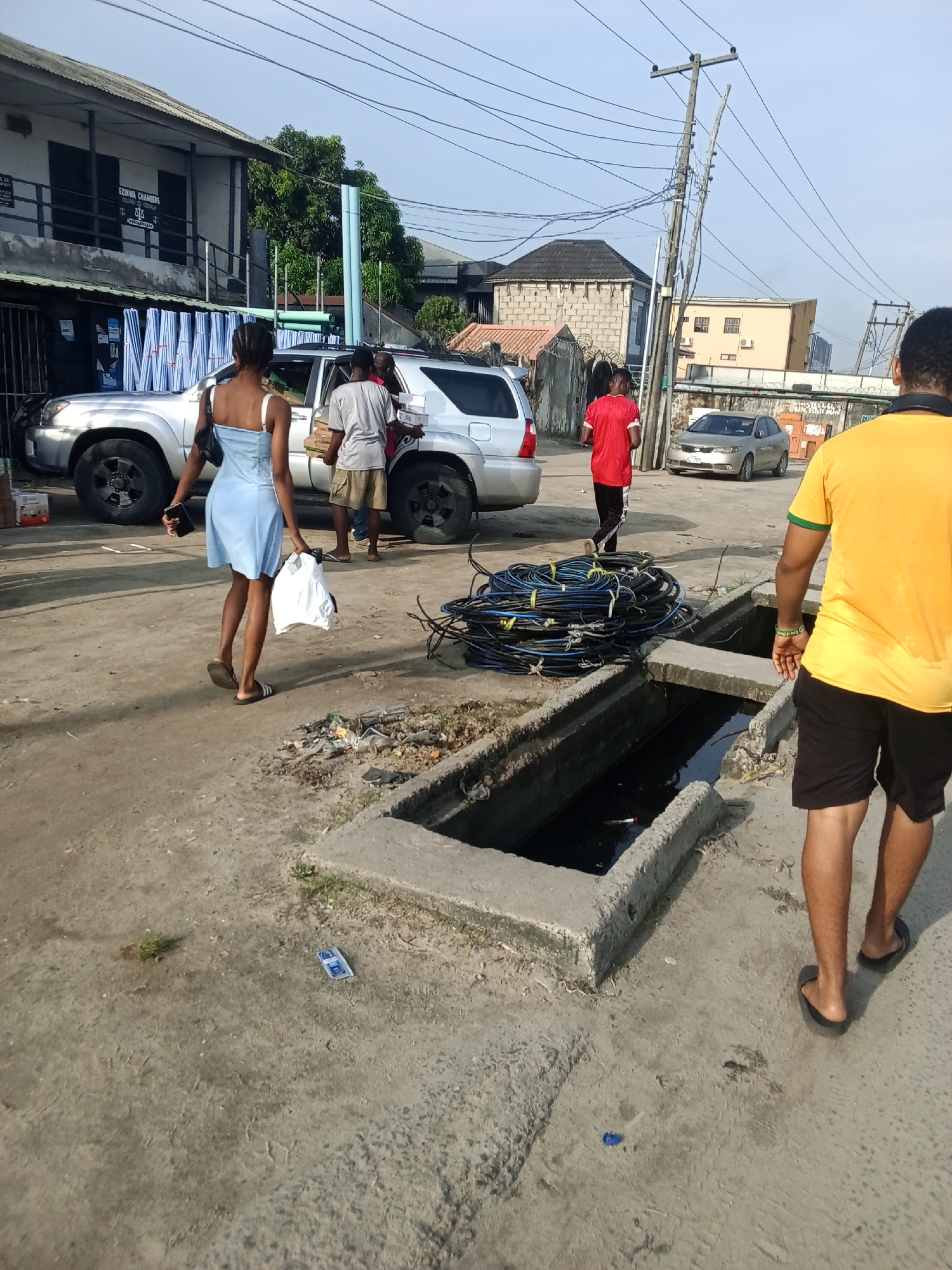 A daytime shot in what appears to be a residential or mixed-use area in Ado, Nigeria. The ground is concrete or asphalt.
A light-colored SUV is parked on the left. People are near it, loading boxes. A woman in a light blue dress walks away from the car, carrying a white bag. Another person in a red shirt is walking down the road away from the SUV. To the right, a man in a yellow shirt and black shorts is walking towards the camera.
In the foreground, there's a large concrete structure, possibly a storm drain or open sewer. A coil of black cables lies near it.
Buildings are visible in the background, and a utility pole with wires is overhead. A parked silver car can be seen down the road.