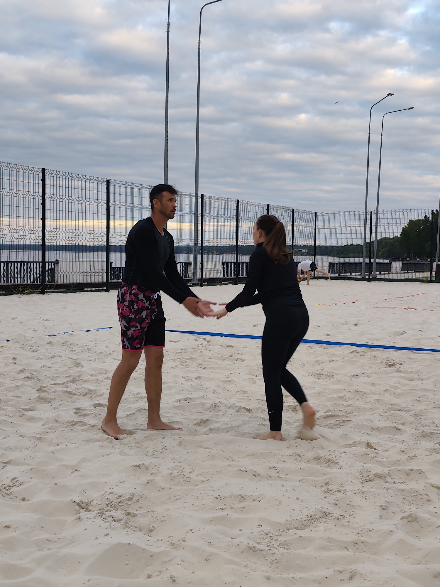 This image depicts a beach volleyball court with two individuals in the foreground engaged in an interaction. A man on the left, wearing a black long-sleeved shirt and patterned shorts, is facing a woman on the right, who is dressed in black leggings and a black long-sleeved top. They are reaching out their hands towards each other, possibly in a gesture of greeting or acknowledgement. The court is covered in sand, with blue and red boundary lines visible. In the background, a chain-link fence separates the court from a body of water, likely a river or lake, with trees and buildings visible on the opposite shore. The sky is overcast with clouds. The scene is set outdoors, with lampposts present on the court. It is plausible this location is the beach volleyball court situated by the Kama River in Chaikovsky, Russia, given the description of the landscape.