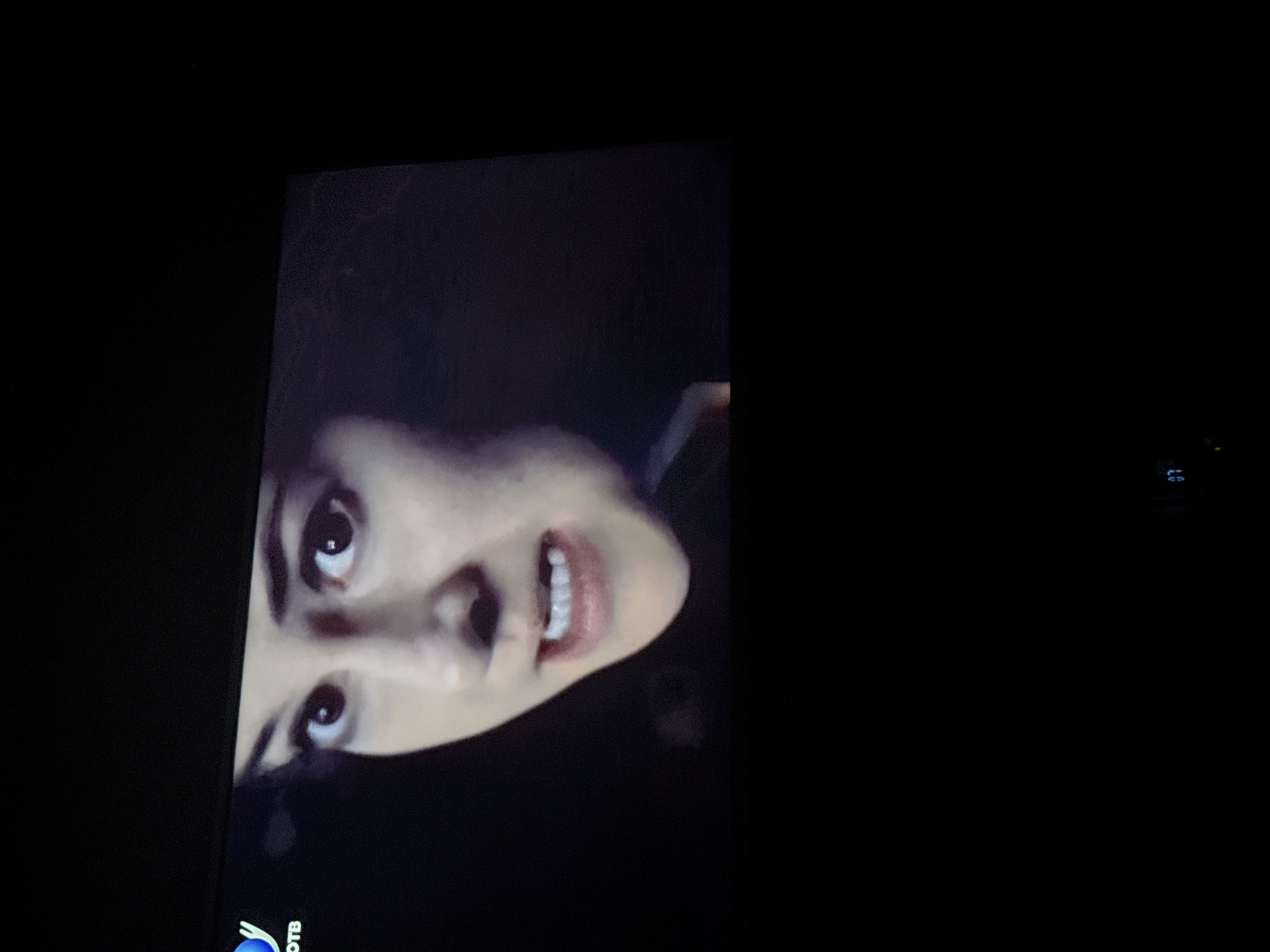 A close-up shot of a person's face is displayed on a screen. The person's eyes are wide, and their mouth is slightly open, revealing their teeth. The background is dark, and there are no other discernible objects or details. The location cannot be determined from the provided image.