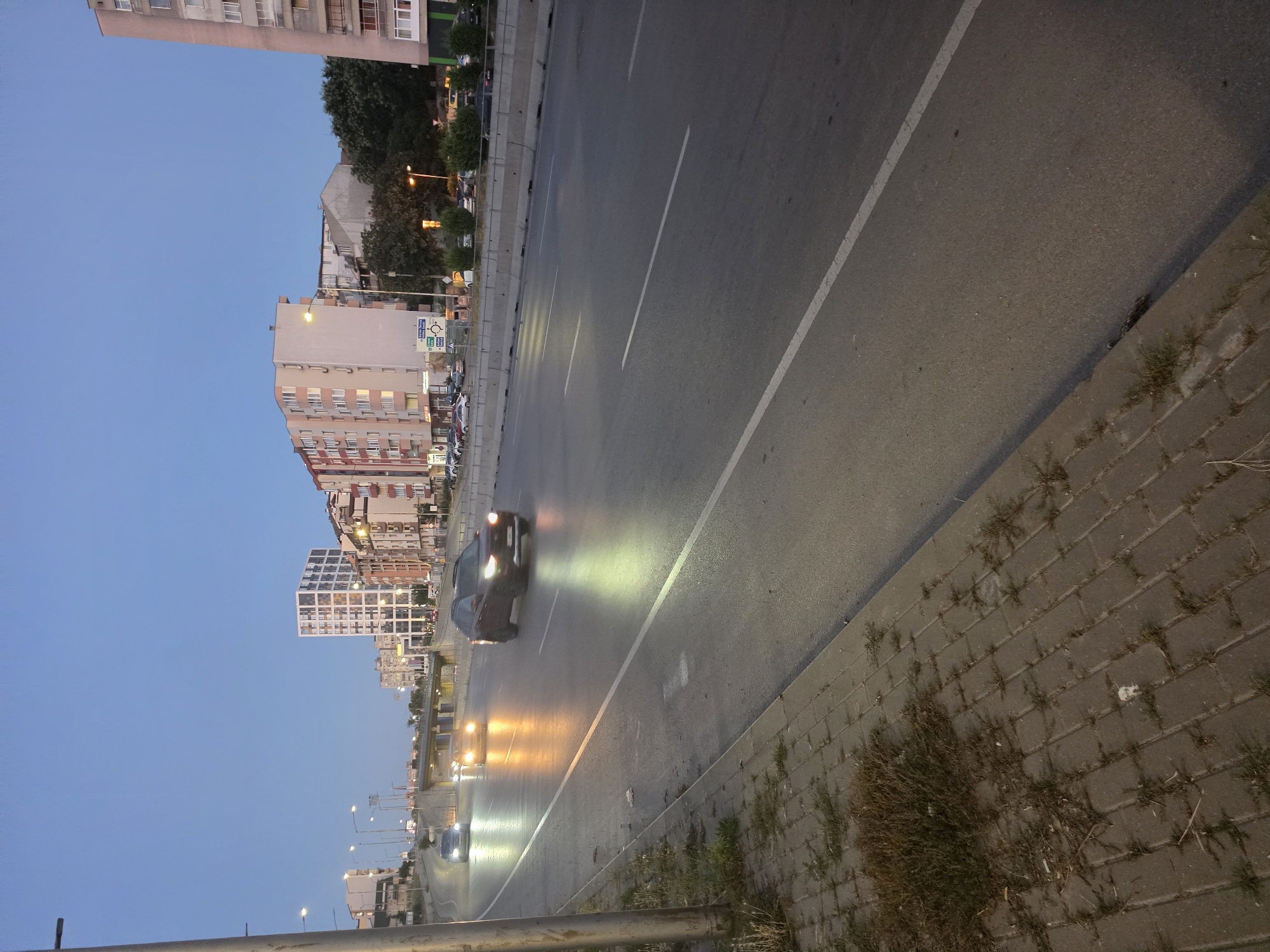 The image depicts a street scene in Varna, Bulgaria, at dusk. The sky is a clear, pale blue, transitioning into twilight. A multi-lane road with visible white lane markings dominates the foreground and midground, with cars driving on it, their headlights illuminated. To the left of the road, a paved sidewalk is visible, with some sparse vegetation growing between the pavers. Beyond the sidewalk, a pole with streetlights stands tall. The background features a cityscape with several apartment buildings of varying heights and architectural styles, some with brightly lit windows, and a modern, white, multi-story building with a distinctive pattern. Trees and other foliage are also present, interspersed with the urban structures. The overall atmosphere suggests a typical urban environment during the evening transition.