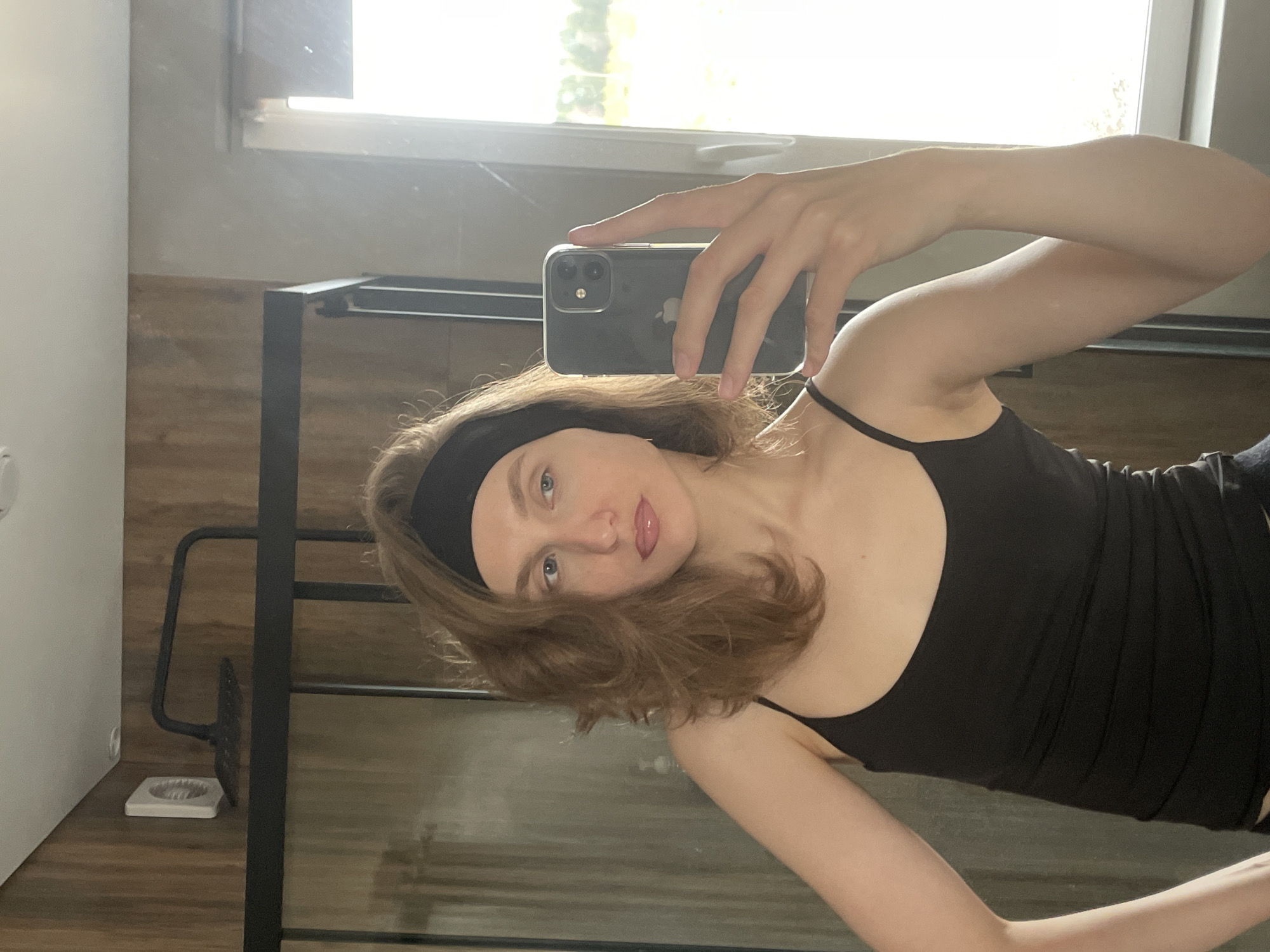 A medium close-up, horizontally oriented mirror selfie features a young woman indoors. She holds a smartphone, specifically an iPhone with a clear case, in her right hand to capture her reflection. The phone's back is visible, showing its dual cameras and the "iPhone" logo.
The woman has light-colored skin, light blue eyes, and shoulder-length wavy brown hair, which is partly held back by a black headband. She is wearing a black spaghetti-strap tank top. Her expression is neutral with a slight pout on her glossy lips. Her body is angled slightly, and her gaze is directed towards the camera.
The background, visible through the mirror, reveals an indoor setting bathed in bright daylight from a large window. Green foliage can be seen faintly outside the window, indicating daytime. The interior features a light-colored wall on the left and a wooden paneling or shelf below the window. A black metal frame structure is prominent in the mid-ground, possibly part of a shelf, table, or furniture. On the wooden surface to the lower left, a small white item, possibly a toothbrush holder or a similar container, is visible next to a metal fixture. The overall impression is a casual, everyday moment captured in what appears to be a modern home environment in Подшивалово, Russia.