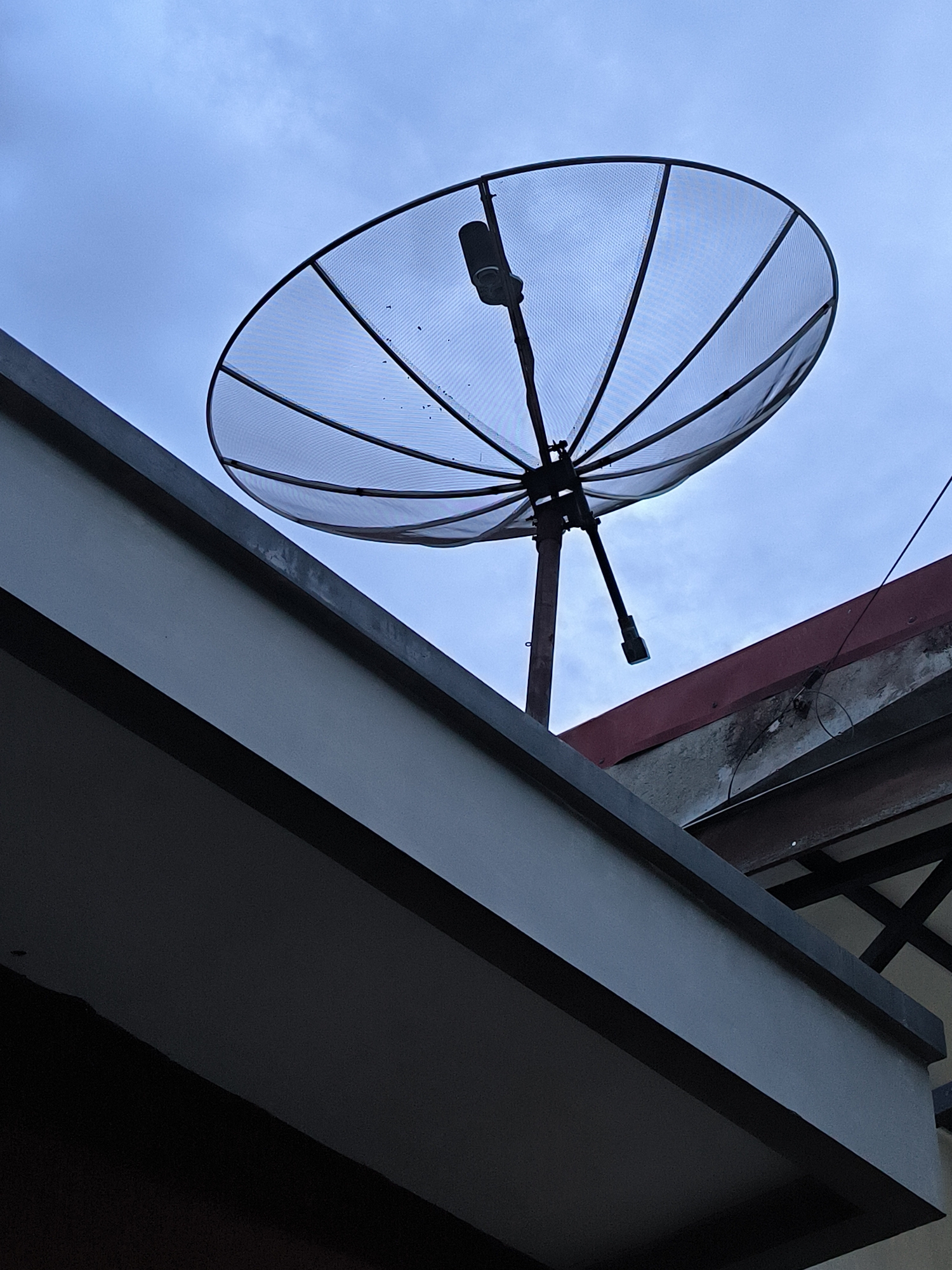 The image shows a satellite dish mounted on a building in Lhokseumawe, Indonesia. The dish is a mesh-type, held by a central support pole. The sky is overcast. Part of the building's structure is visible in the foreground, along with another section of roof behind the dish.