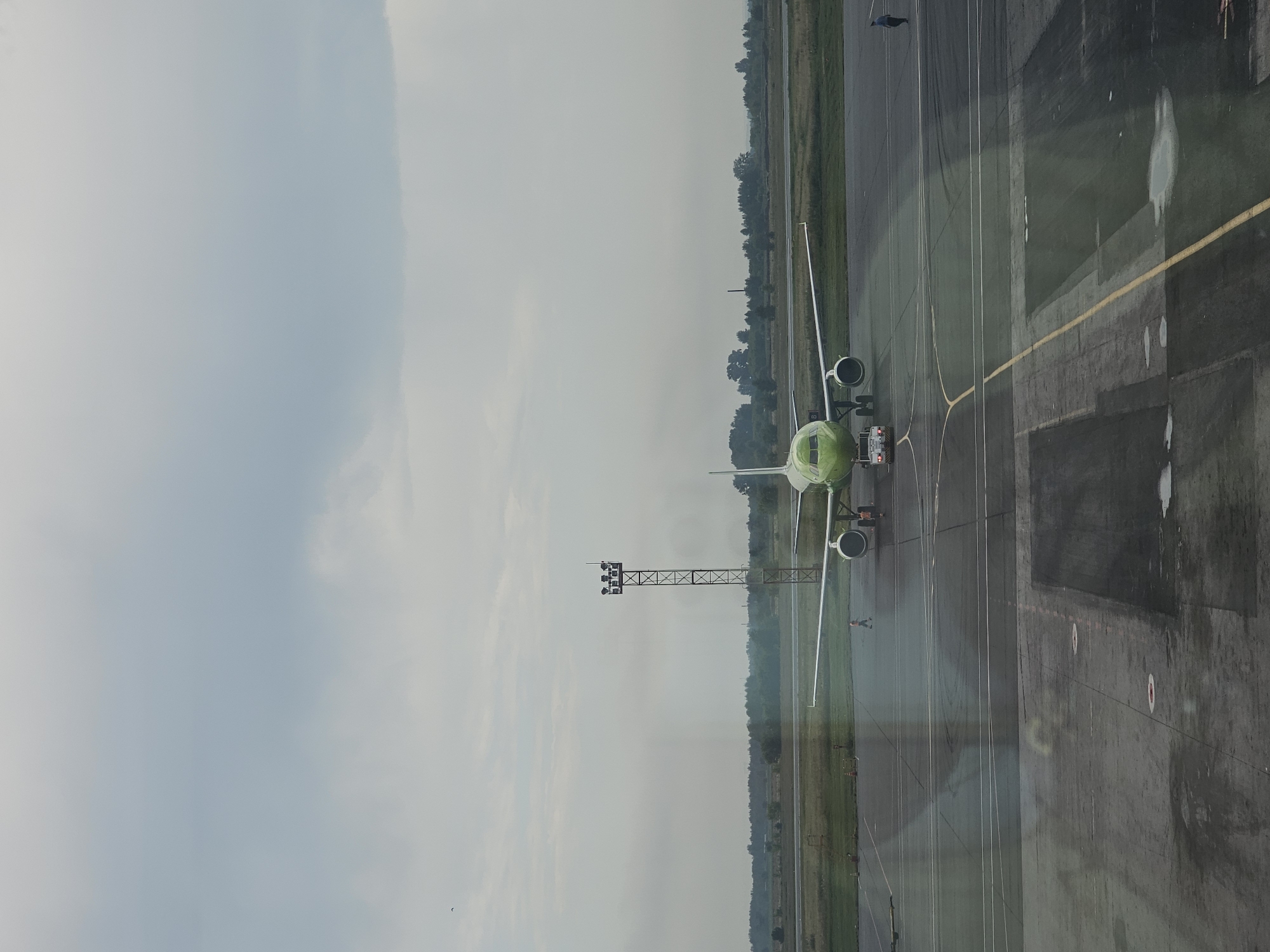 An overhead view of a green airplane parked on the tarmac at Tolmachevo Airport in the city of Ob, Russia. The airplane is positioned in the center of the frame, facing the camera. The sky is overcast with a pale blue hue. In the background, a treeline is visible, with a tall metal tower with lights situated to the left of the airplane. Several people are present on the tarmac, appearing to be airport personnel. The tarmac is wet and reflects the airplane.