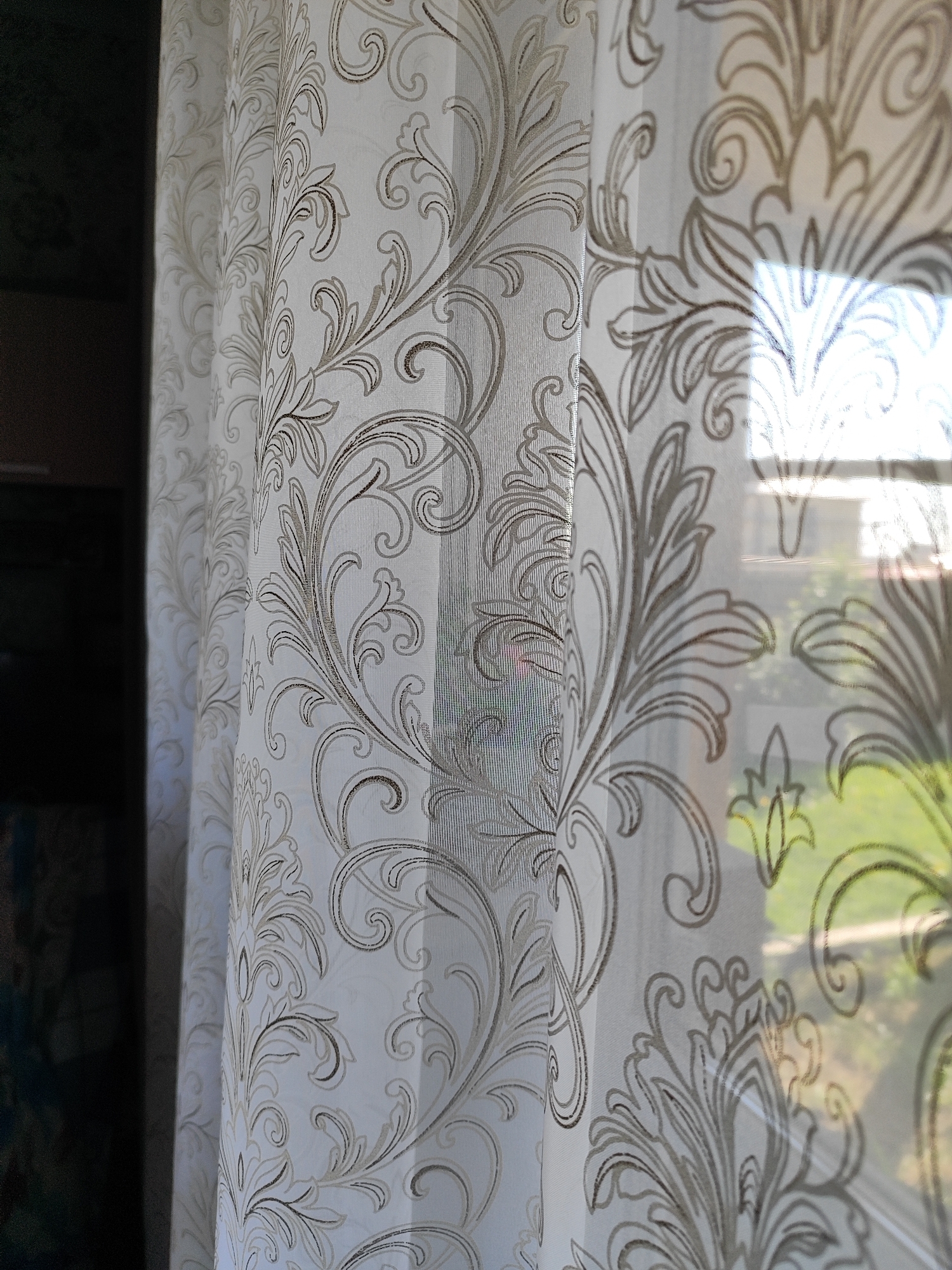 A close-up, eye-level shot shows a white sheer curtain with a repeating damask-like pattern of scrolling vines in beige and taupe. The curtain is semi-transparent, revealing a blurred view of a window with sunlight streaming through. The sunlight creates bright rectangular highlights on the curtain. On the left side of the image, a darker object, possibly a wall or another curtain, is visible, providing a contrast to the lighter fabric. The scene is indoors, in Marusino, Russia. There are no people or actions depicted in the image.