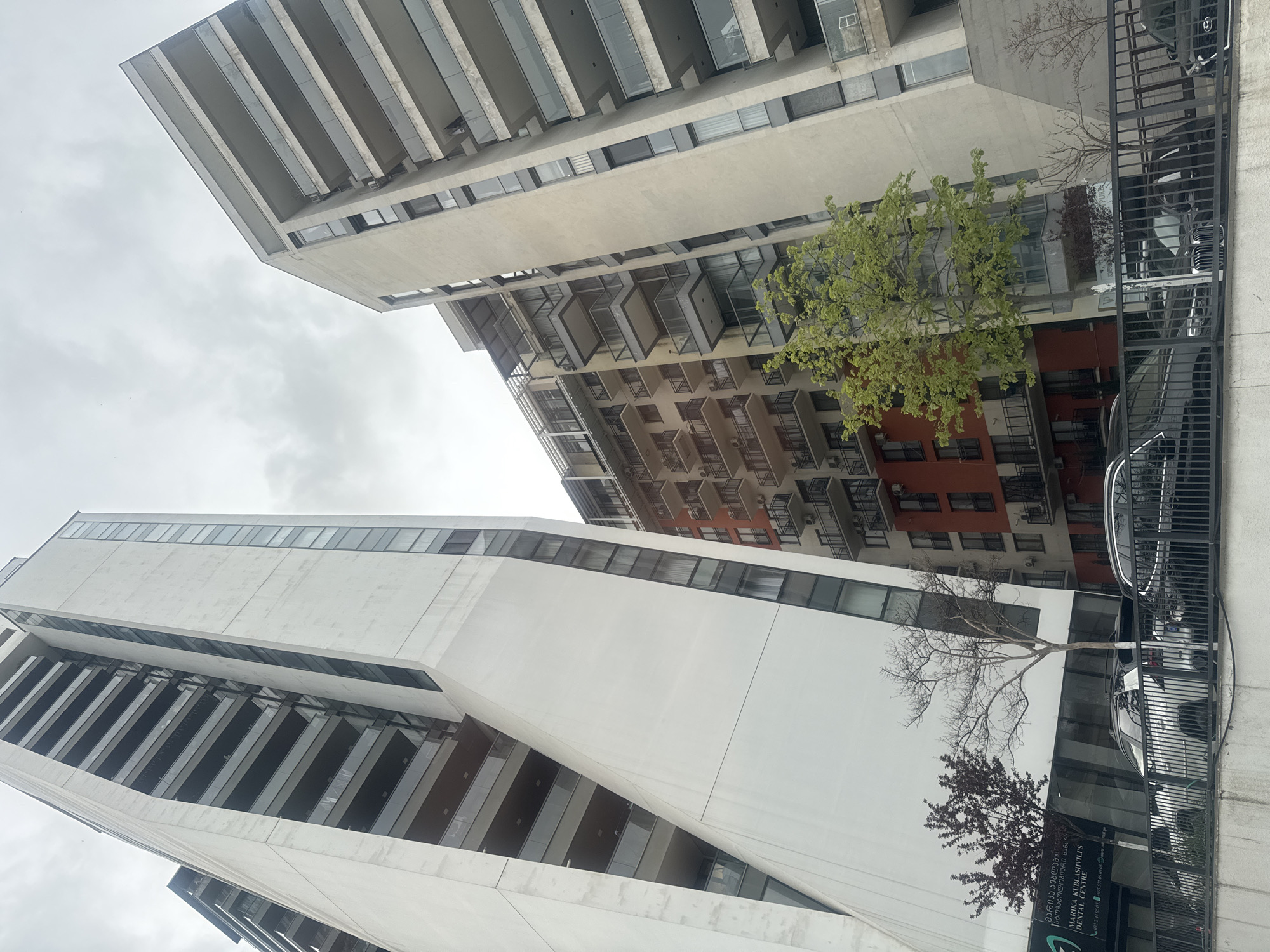 The image presents an urban scene in Tbilisi, Georgia, featuring several modern high-rise buildings under an overcast sky. On the left, a striking white building exhibits contemporary architecture with sharp geometric angles, contrasting dark-paned windows, and integrated balconies. Behind it and slightly to the right, another multi-story building features a lighter beige upper section with numerous open balconies, transitioning to a distinct reddish-brown facade in its middle section, also with balconies. A third building is partially visible further in the background.
At street level, a black metal fence separates the buildings from a street or parking area where parts of dark-colored vehicles are discernible. Prominently in the mid-ground, a vibrant green tree adds a natural element to the concrete landscape. Closer to the ground, a bare, skeletal tree and another with reddish foliage are also present. The sky is uniformly light grey and cloudy, indicating an overcast daytime.
A notable detail is a sign on the ground floor of the white building, clearly displaying "MARIKA KURIASHVILIS DENTAL CENTRE" in English, accompanied by Georgian script and contact information.