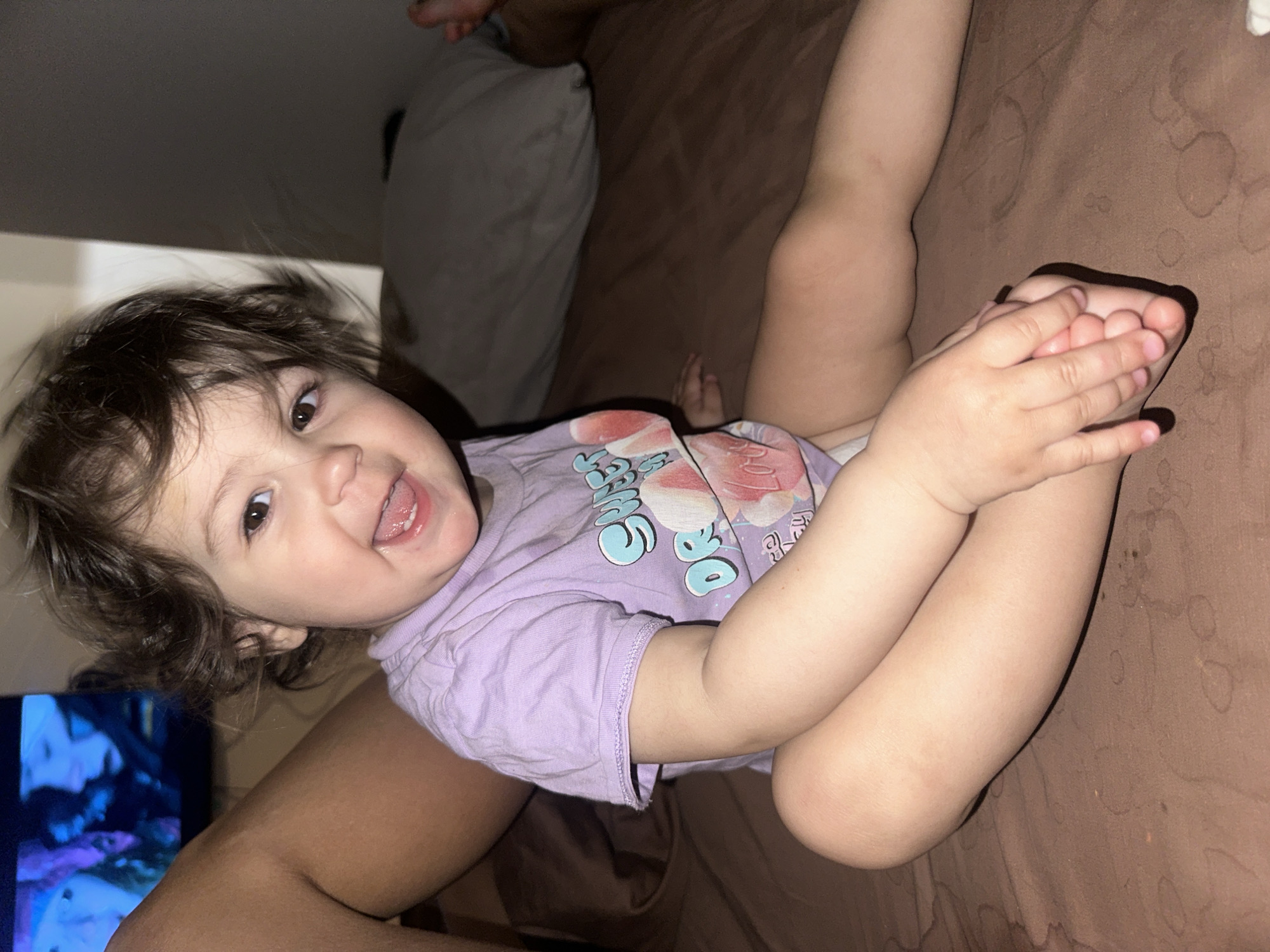 A toddler with curly brown hair and brown eyes, wearing a light purple t-shirt with a graphic design, is lying on a brown bed. The toddler is smiling and holding their feet with their hands. In the background, there is a pillow and a leg of another person. A television is visible in the lower left corner of the image. The location is unknown, and there is no indication that the image was taken in Khimki, Russia.