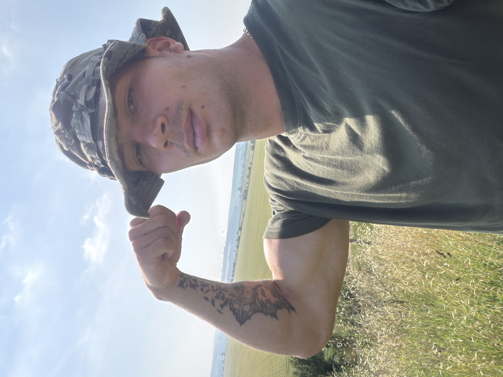 A male individual, appearing to be in his late 20s to early 30s, stands outdoors in Bogdanitsa, Bulgaria, under a bright, clear sky with scattered cirrus clouds. He wears a camouflage boonie hat and a plain olive green short-sleeved t-shirt. He is positioned facing the camera, flexing his right bicep, prominently displaying a large, intricate tattoo on his right forearm that features a dark, stylized bat-like creature with orange accents and abstract dark elements extending up his arm. His expression is neutral as he looks directly into the lens. In the immediate foreground, tall, light-green and golden grasses are visible. The midground and background consist of expansive, verdant agricultural fields stretching across gently rolling terrain under full sunlight. A faint, slender structure, possibly a tower, is discernible on the distant horizon.