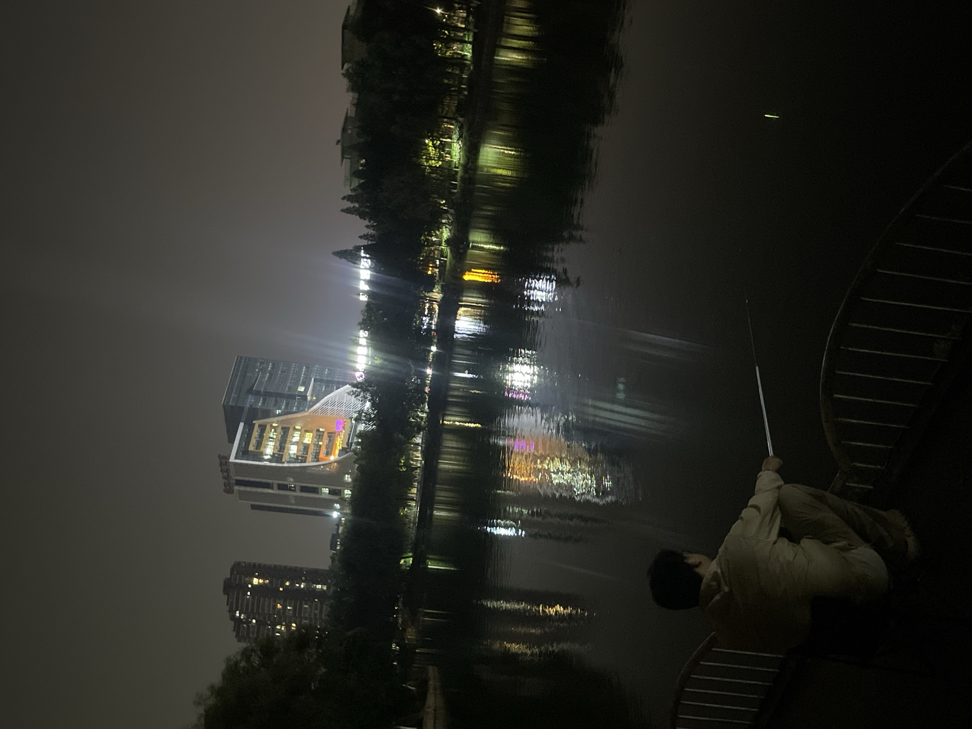 This nighttime photograph, oriented vertically, captures a serene scene of a person fishing by a body of water in an urban environment. The sky is dark and appears to be overcast.
In the immediate foreground, a person, likely male, is seated or crouching on a dark, curved metal railing. He is wearing light-colored, possibly beige, clothing and holds a long fishing rod that extends out over the water. The person's back is to the viewer, focused on the activity.
The body of water, likely a river or lake, fills the midground, acting as a canvas for the vibrant reflections of distant lights. These reflections appear as elongated, shimmering streaks of various colors—yellow, orange, white, and hints of purple—indicating light sources from the opposite bank. A small, distinct green light is visible on the water's surface further out, possibly a fishing float.
Across the water, several modern buildings stand tall against the dark sky. Their windows and facades are brightly illuminated, casting a significant glow that is diffused by the night air. Trees with dense foliage line the bank opposite the viewer, partially obscuring some of the structures but also reflecting their own lit-up greenery in the water.
The overall impression is one of quiet contemplation amidst an urban backdrop, with the solitary act of fishing contrasting with the illuminated city lights. There is no visible text in the image.