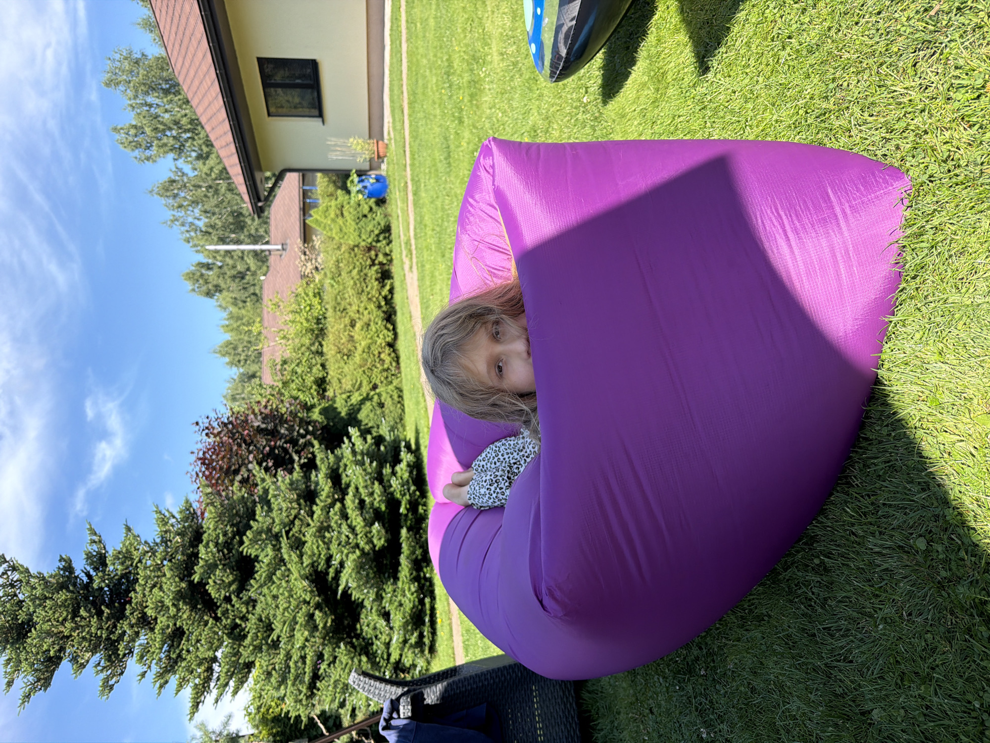 A young girl with blonde hair and a white shirt with a leopard print is resting inside a purple inflatable lounger in a grassy backyard. She is peeking over the edge of the lounger, looking at the camera. The lounger is positioned on a well-maintained green lawn. In the background, there is a large evergreen tree, a red-leafed bush, and a light-colored house with a brown roof. The sky is blue with some white clouds. There is also a wicker chair to the left of the frame and a portion of another inflatable object to the right. This scene is set in the city of Baloži, Latvia, on a sunny day.