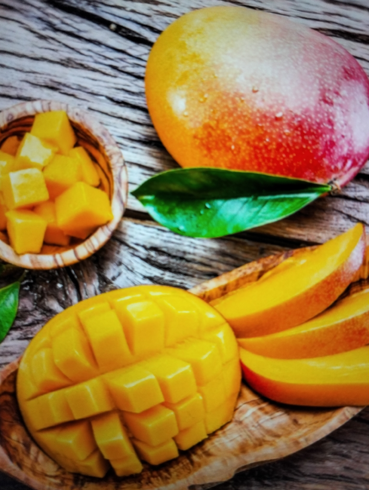 The image shows a rustic wooden surface with several mangoes. In the upper right is a whole mango, transitioning from yellow to orange and red, with small water droplets on its skin. To its left, a small wooden bowl is filled with cubed mango. Below these, a halved mango, scored into a grid pattern, rests on a wooden platter. To the right of the halved mango are three sliced mangoes, fanned out. Two green mango leaves are also visible, one near the whole mango and the other near the bottom left corner. The scene is lit from above, highlighting the textures of the mangoes and the wood. There are no people or specific landmarks indicating a location in Payagaboh, Indonesia.