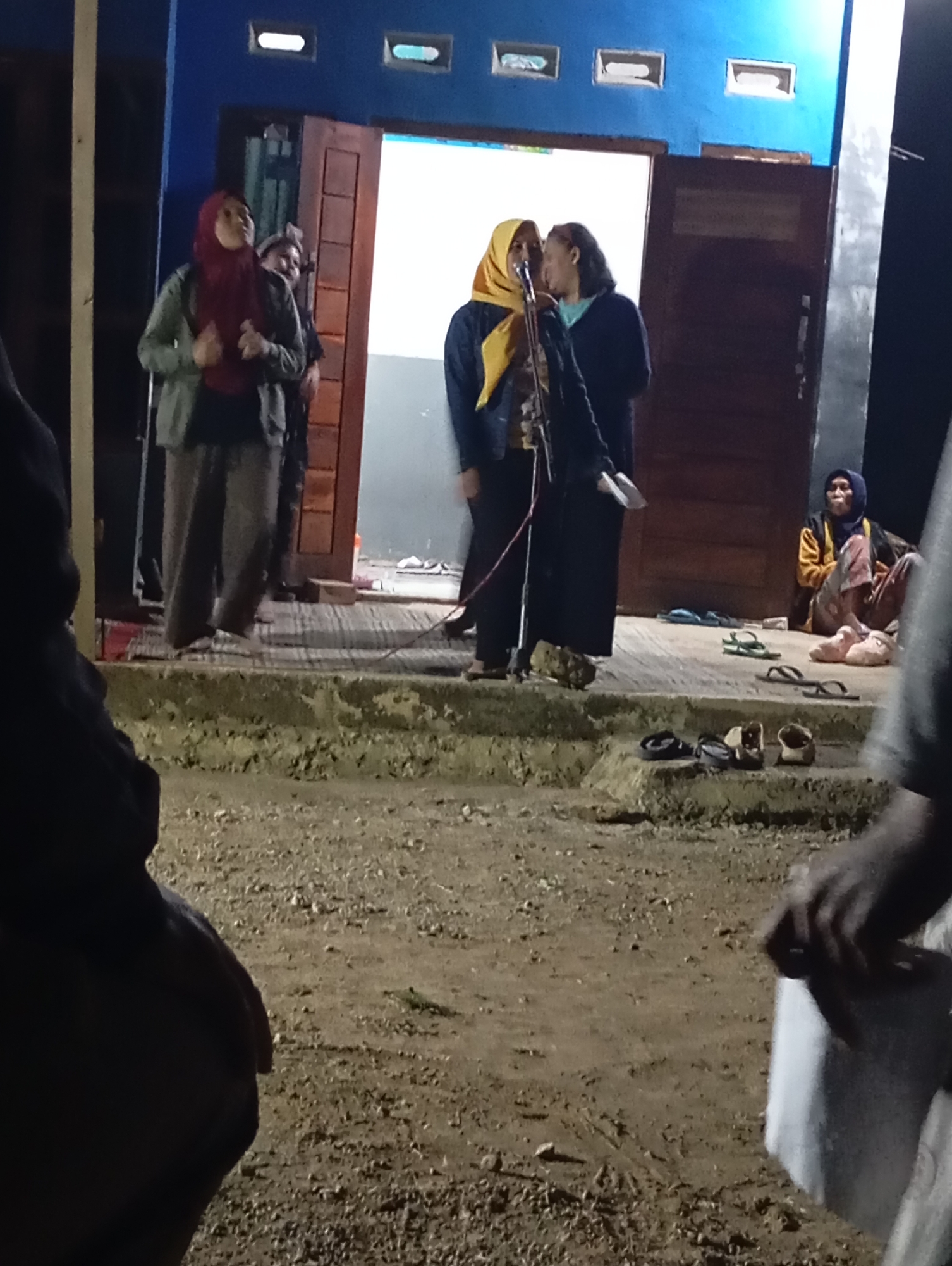 In Ketepung, Indonesia, a group of people are gathered outside a building at night. Two women are standing near a doorway, one speaking into a microphone. The woman on the left wears a red hijab and a light-colored jacket over a dark shirt, with baggy pants. The woman on the right has a yellow hijab and a dark jacket over a lighter shirt. Behind them, another woman with dark hair and a blue shirt is partially visible. To the right, a woman wearing a blue hijab, an orange jacket, and patterned pants sits on the ground. Several pairs of sandals are scattered on the ground and steps in front of the building. The ground is covered with dirt and small stones, and there is a concrete step. The building has a blue facade with two rectangular windows and a dark wooden door.