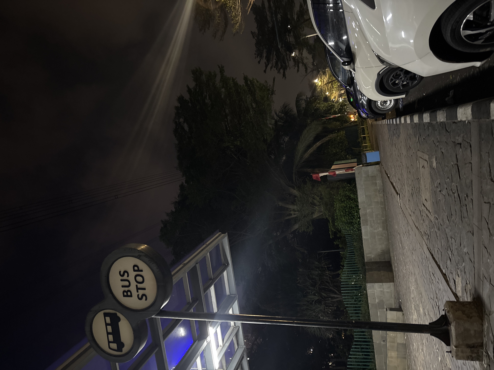 At night, a bus stop sign with a bus silhouette and the words "BUS STOP" is illuminated. To the right, three cars are parked: a dark-colored car, a white car, and another white car in the foreground. The ground is paved with cobblestones, and a green fence is visible in the background. The scene appears to be in North Jakarta, Indonesia, based on the overall environment.