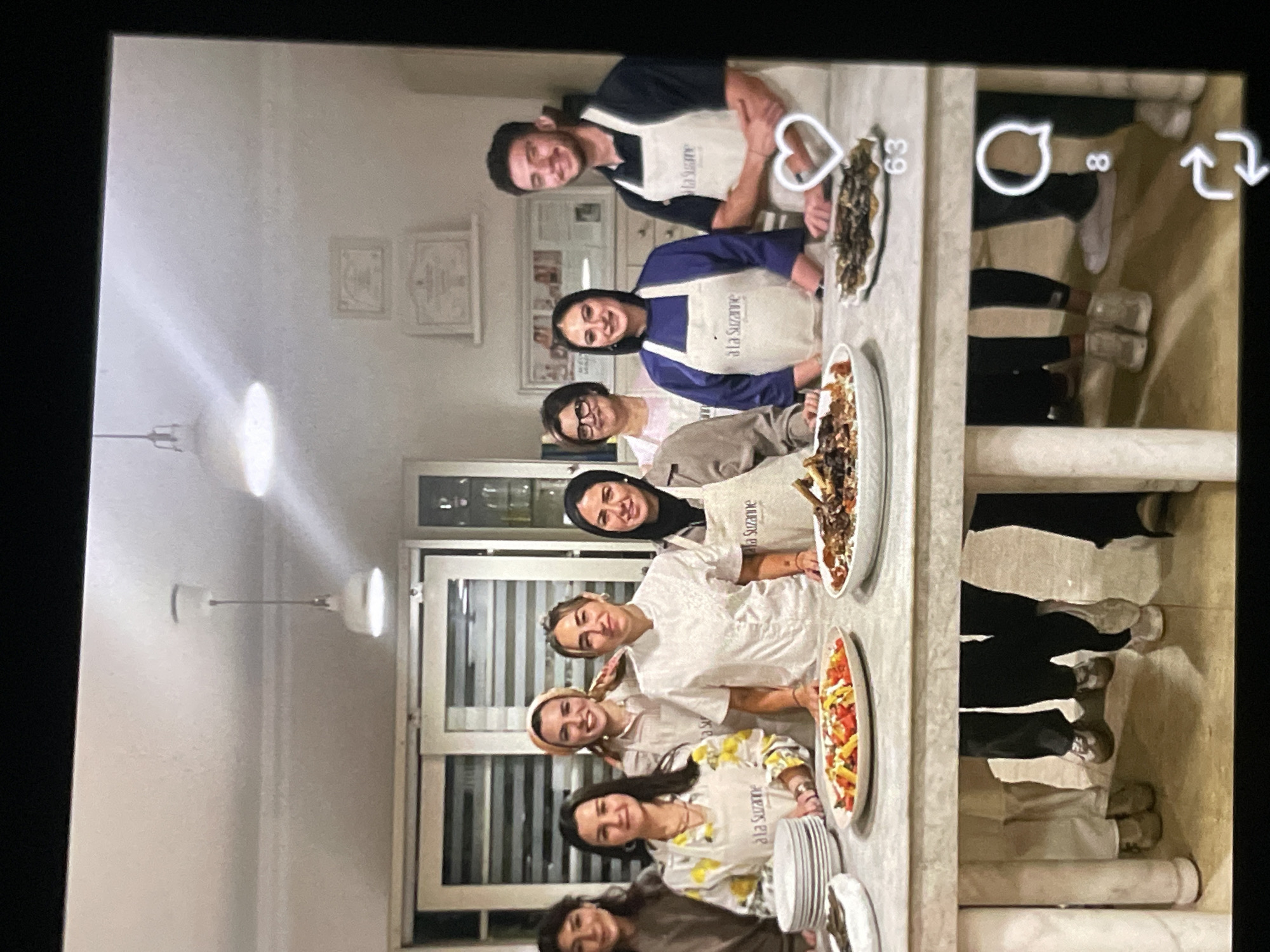 A group of eight people, six women and two men, stand behind a table in a kitchen. The people are all wearing aprons, and some have chef hats or headscarves. The table is laden with food, including platters of meat and a salad. In the background, there are kitchen cabinets and a refrigerator. The scene appears to be a cooking class or demonstration. The location is identified as Unknown, Unknown.