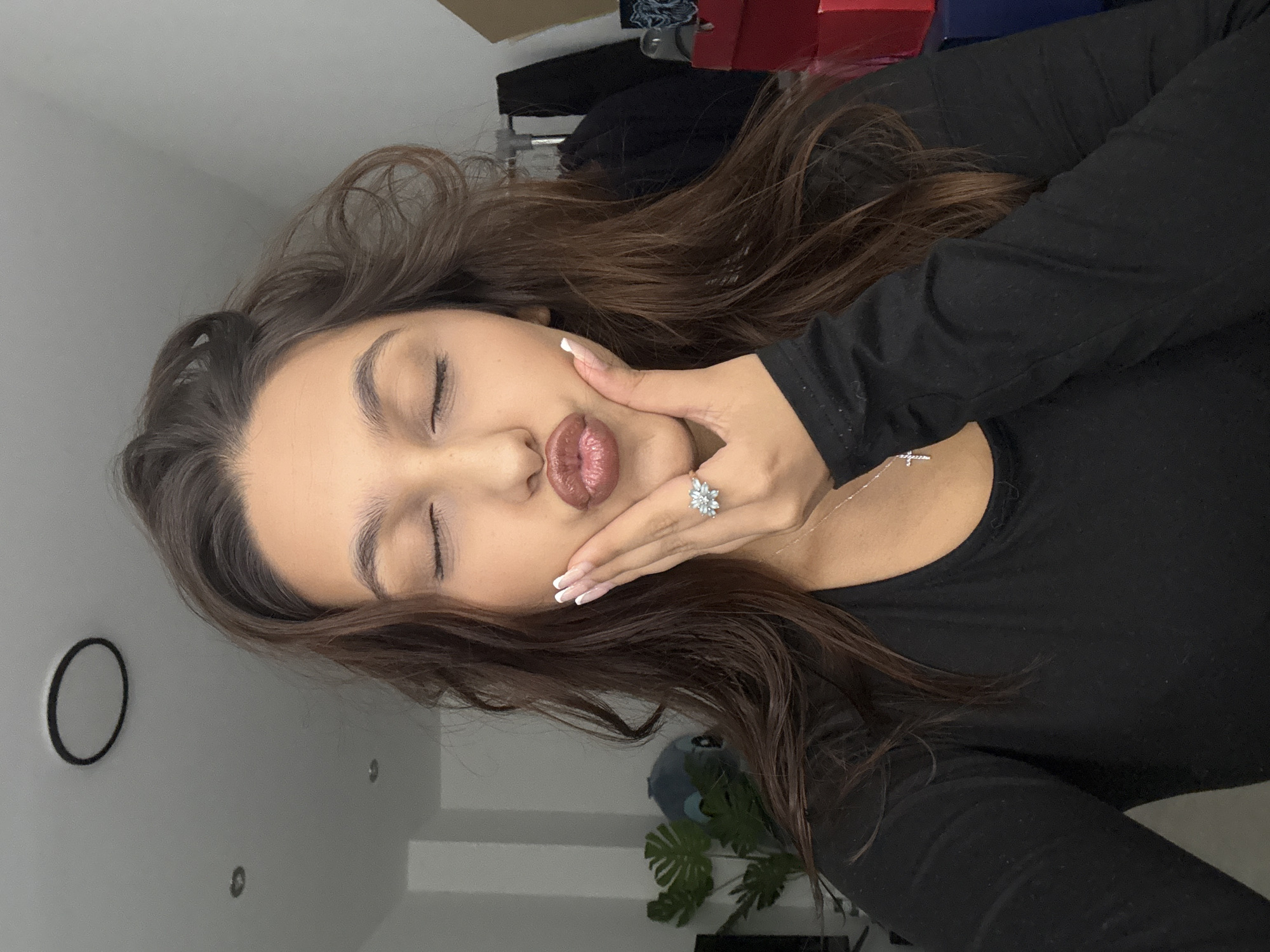 The image displays a close-up portrait of a young woman posing indoors in Mönchengladbach, Germany. She has long, wavy dark brown hair framing her face. Her eyes are closed, and her lips are pursed in a "kiss" expression, highlighted with glossy brownish-red lipstick. Her right hand, adorned with a silver-toned ring featuring a floral or starburst design on her ring finger and manicured nails with white tips, rests gently against her cheek. She is wearing a plain black long-sleeved top and a delicate silver necklace with a small cross pendant.
The background is softly blurred but reveals elements of an indoor setting, likely a room within a home. A light-colored wall is visible, along with an artificial plant featuring broad green leaves. Other indistinct objects, including dark furniture or boxes and possibly some red boxes, can be seen. A black circular object is mounted on the wall in the upper left section of the frame. The lighting is soft and even, suggesting an indoor setting without direct harsh light. The woman's pose conveys a playful and relaxed demeanor. There is no visible text in the image.