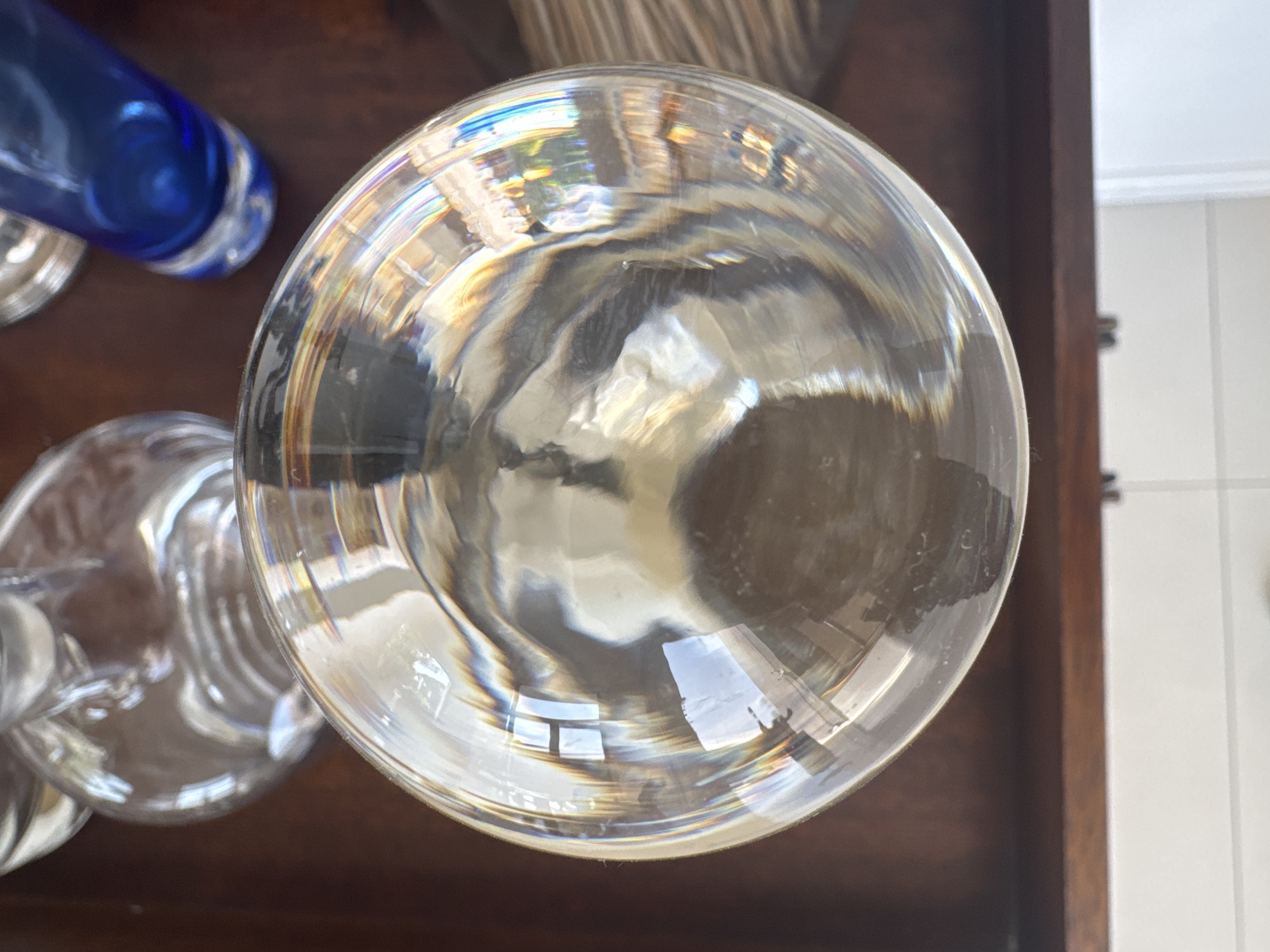 The image shows a close-up, overhead view of a collection of glassware sitting on a dark brown, possibly wooden, surface. Predominantly in focus is a large, clear glass goblet or bowl, its circular shape dominating the center of the frame. The glass refracts light, creating a swirling, distorted view of the surroundings. To the left of the large glass, other pieces of glassware are partially visible, including a blue glass object and another clear glass with a stem. The surface beneath the glasses appears to be part of a drawer or cabinet. A portion of a white wall or tile is visible in the upper right corner of the frame. The location and specific details related to Puebla, Mexico, cannot be determined from the image itself.