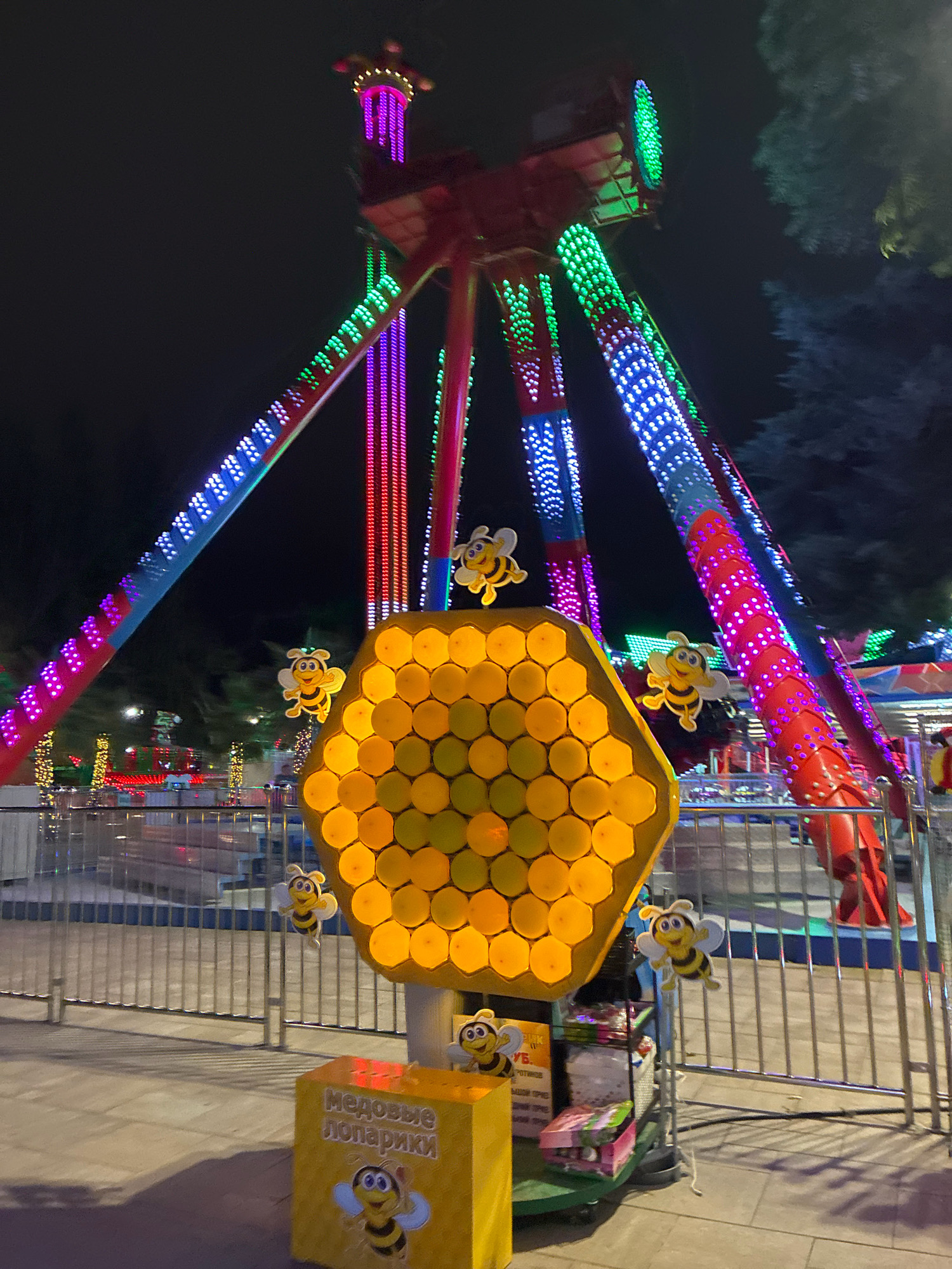 An outdoor amusement park scene at night in Sochi, Russia, features a large, red swing ride with multiple arms extending upwards, adorned with colorful LED lights in pink, green, and blue. In the foreground, a large, hexagonal, honeycomb-shaped structure glows with warm yellow light, decorated with cartoon bee figures. A yellow, cubic kiosk with Russian text, likely indicating a game or attraction, is positioned in front of the honeycomb. The area is enclosed by a metal fence, and blurred lights and trees are visible in the background. The ground is paved.