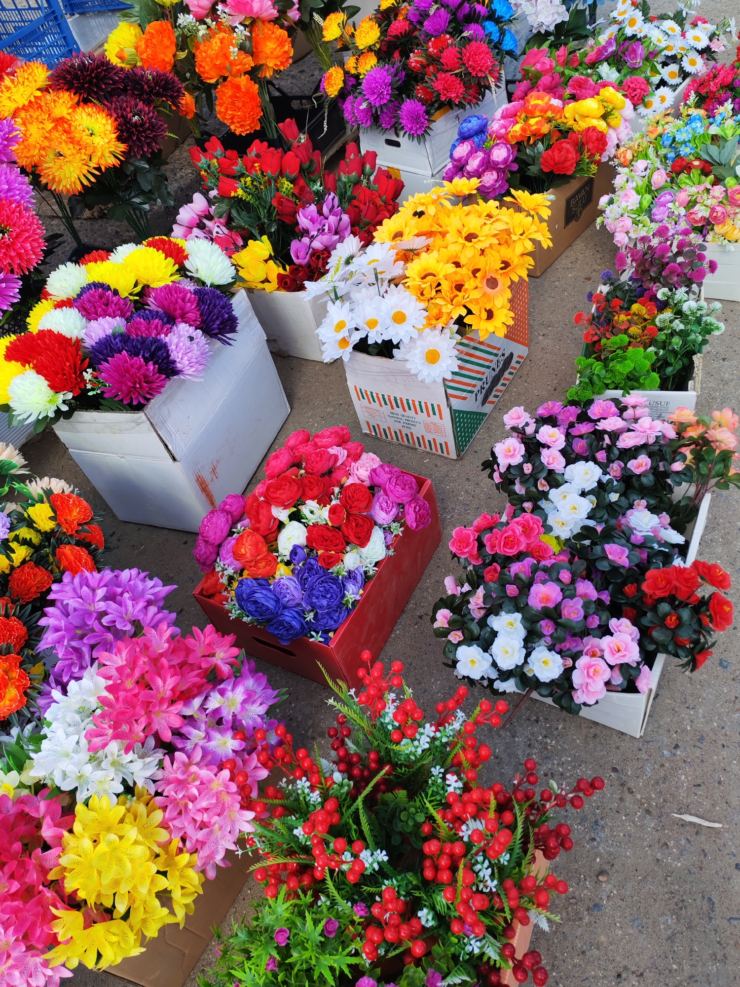 This high-angle shot captures a vibrant and extensive display of artificial flowers, likely set up at an outdoor market or street vendor stall in Chaikovsky, Russia. The scene is brimming with densely packed bunches of colorful faux blossoms, arranged in various cardboard boxes and plastic crates on a concrete ground surface.
The array of artificial flowers is diverse, featuring types resembling chrysanthemums, daisies, roses, lilies, and small red berries with green foliage. The color palette is exceptionally broad and vivid, encompassing bright reds, oranges, yellows, greens, blues, purples, pinks, fuchsias, and whites. Each container often holds a specific type or color arrangement, creating distinct clusters of color throughout the frame. For instance, some boxes feature large, multi-petaled blooms in purples and reds, while others contain bright yellow daisies or realistic-looking red berry branches.
The arrangement suggests a commercial setting where these flowers are sold, indicating an active trade or market event. The bright and even illumination points to daytime. No individuals are visible in the image, allowing the focus to remain entirely on the colorful merchandise.
Visible text on some of the packaging includes branding and product information. A striped cardboard box in the lower left section clearly reads "HIGH QUALITY PRODUCTS FOR EXPORT" and "PRODUCT OF TURKEY," along with "PROLES." Another brown box shows partially visible text that appears to be "BEYAN SAVUN," while a white box on the right has "YUSUF" printed on it. The overall impression is one of abundance and cheerful artificial beauty, catering to decorative needs.