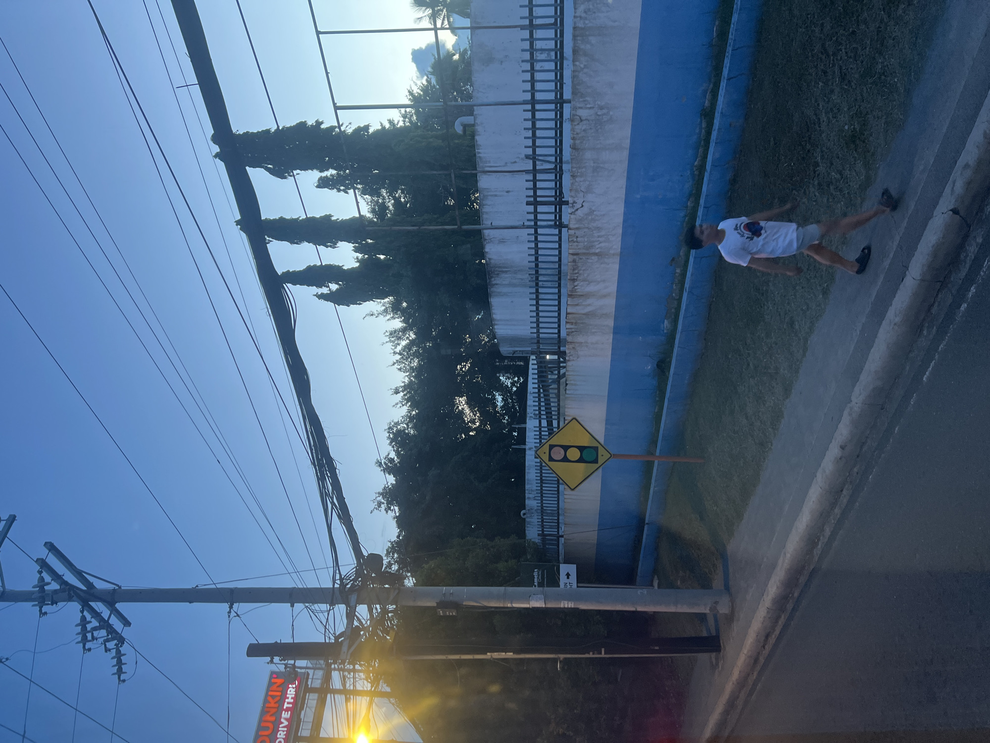A young man wearing a white t-shirt and light-colored shorts walks along a sidewalk next to a road. He is on the right side of the frame, heading away from the viewer. Behind him, a yellow diamond-shaped traffic sign with a pedestrian symbol is visible. The sidewalk is bordered by a grassy area and a blue and white concrete wall with a black fence on top. In the background, tall trees and utility poles with numerous wires are seen against a clear blue sky. A sign for "Dunkin" is visible on the left side of the image. The scene appears to be in an urban environment in Davao City, Philippines, possibly near a commercial establishment. The lighting suggests it is either early morning or late evening.