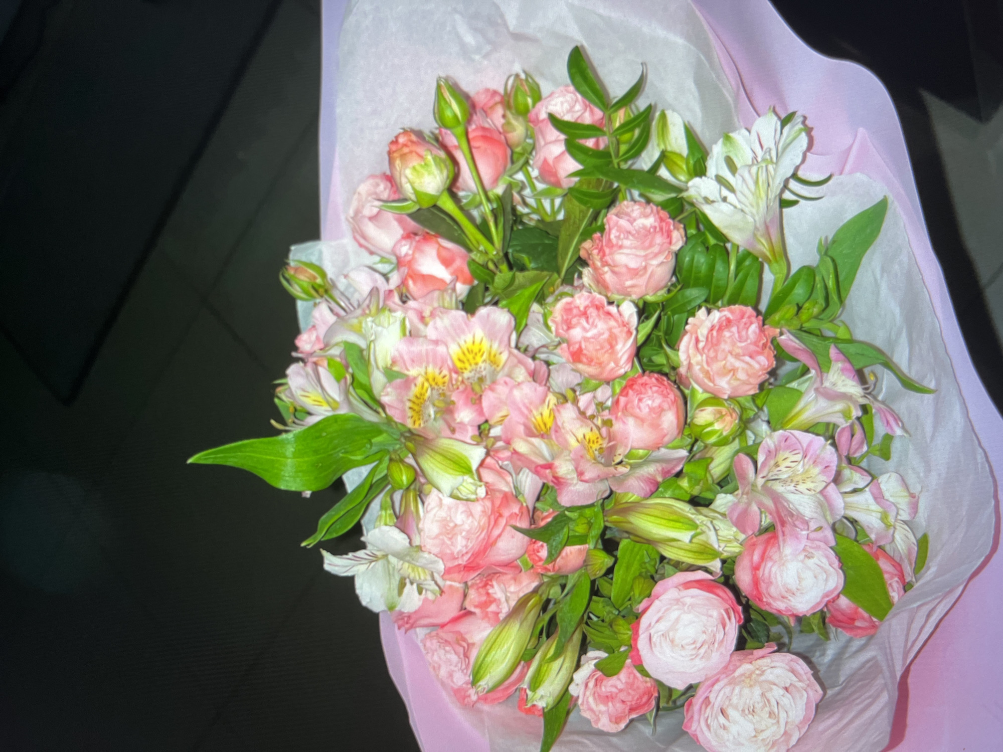 A bouquet of pink roses and white alstroemeria flowers is presented in a light pink paper wrapping. The bouquet is arranged in a dense cluster, with some roses in full bloom and others as buds. Green leaves and stems are visible throughout the arrangement. The background is dark and out of focus, suggesting an indoor setting with low lighting. There are no people or animals visible in the image. The image does not provide any information about its location in Dubai, United Arab Emirates.
