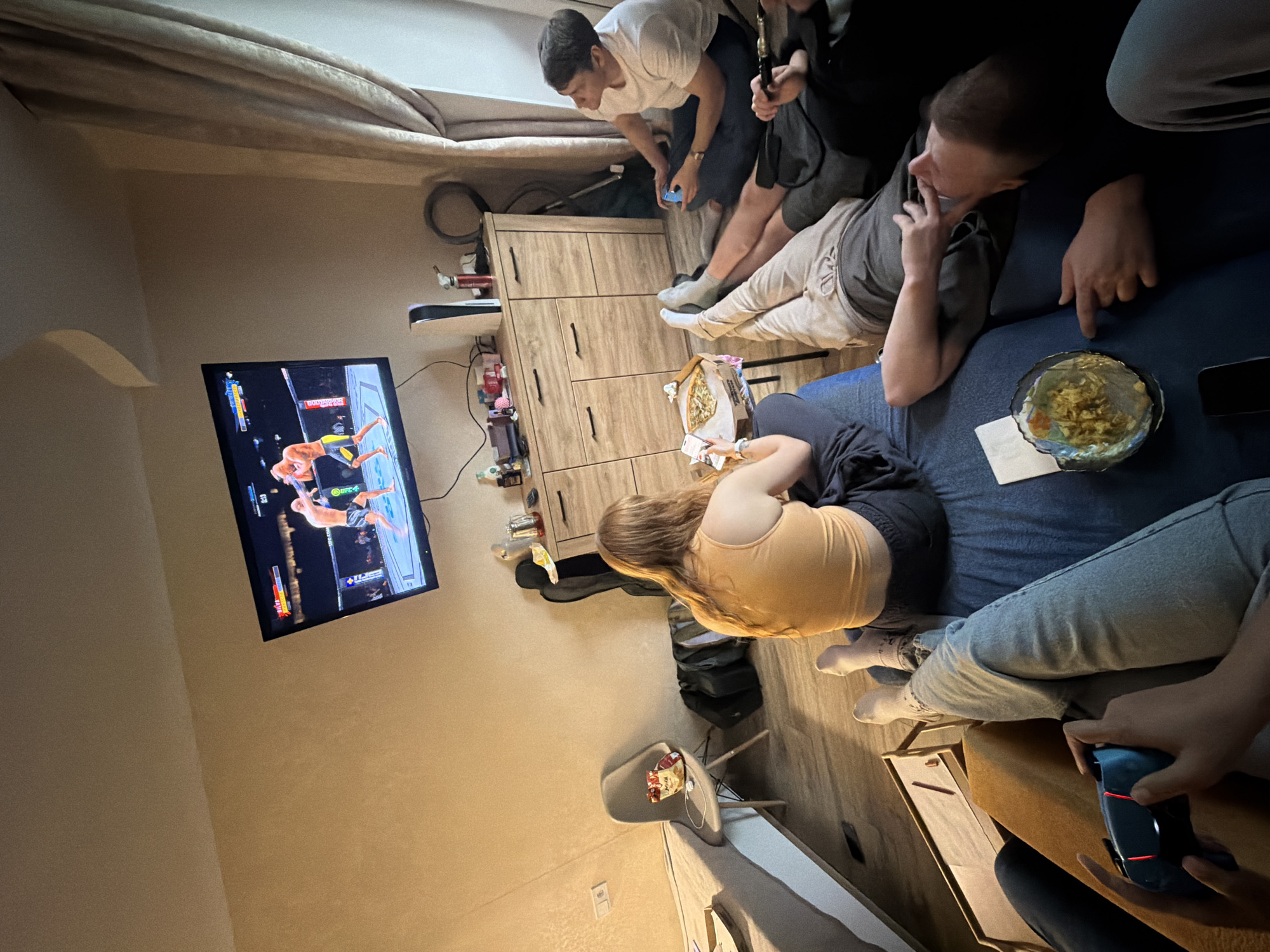 A group of people are gathered in a room, watching a mixed martial arts event on a TV mounted on the wall. Some are seated on a blue sofa. There is a bowl of food on the sofa, and people are sitting nearby. A man is bending over a wooden dresser while holding a video game controller. A chair is located near the TV. The location is in the city of Unknown, Unknown.