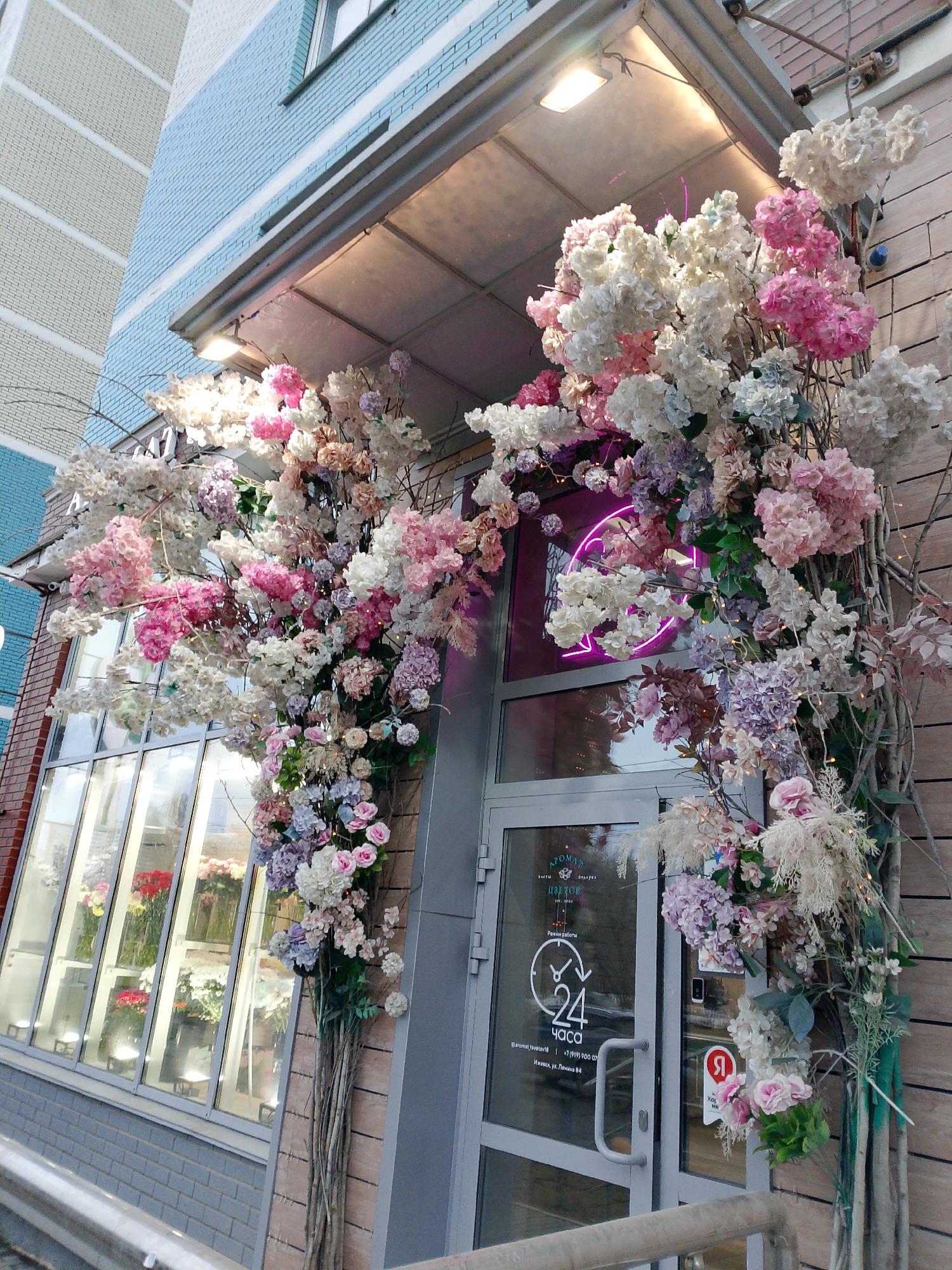 Here is a description of the image:
The exterior of a flower shop called "Aromat Tsvetov" is lavishly decorated with pink, white, and purple flowers forming an arch around the entrance. The shop's glass door has a sign indicating it is open 24 hours. Inside the shop, various flowers are visible. The shop is located in Izhevsk, Russia, specifically on Lenin Street, number 84. The shop's building has a light blue and grey exterior with a light overhead covering the entrance.
