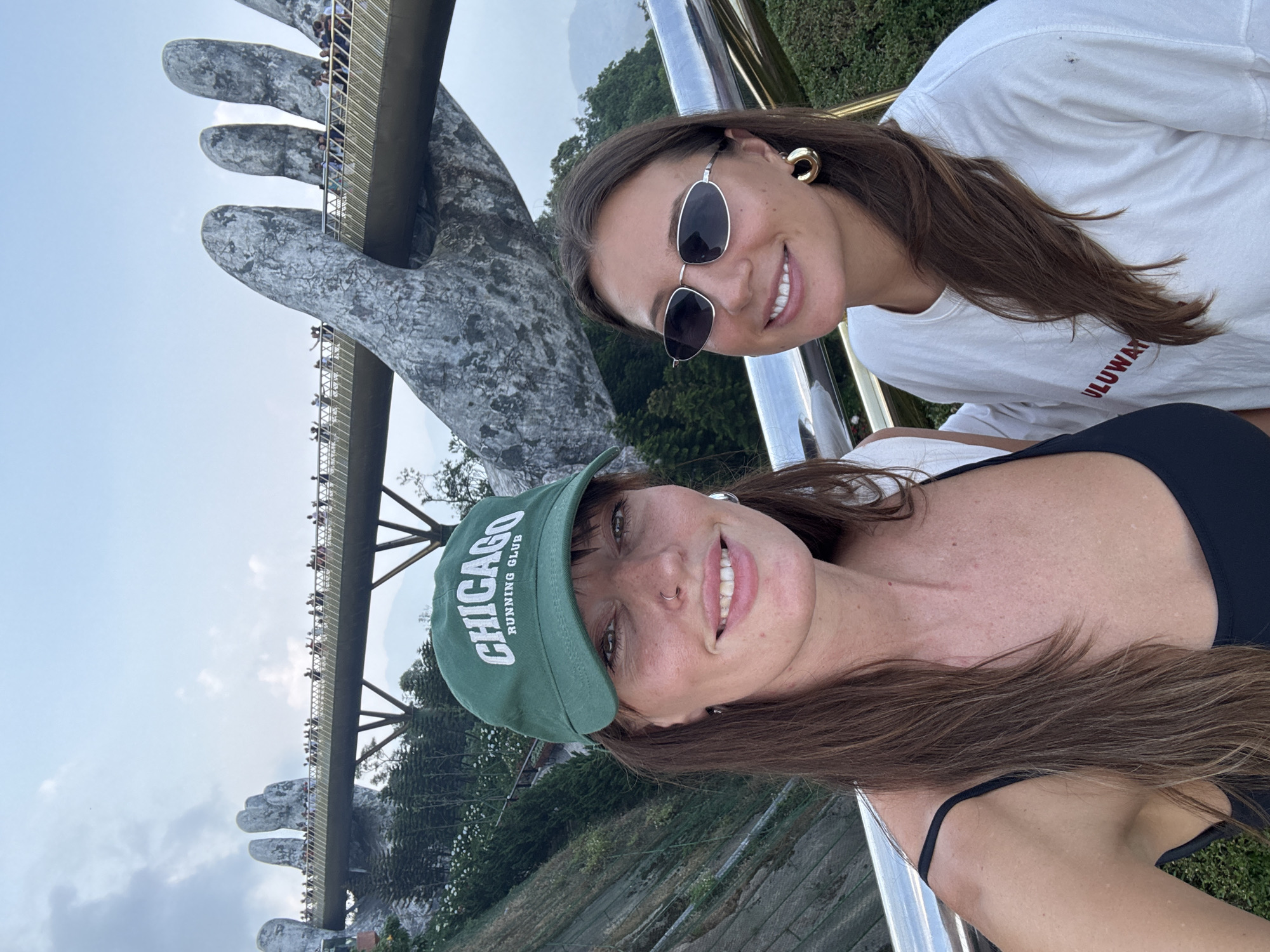 Two smiling women are taking a selfie with the Golden Bridge in Đà Nẵng, Vietnam, as the backdrop. The Golden Bridge is held up by two giant stone hands and is filled with people. The woman on the left is wearing a green "Chicago Running Club" baseball cap and a black tank top, while the woman on the right is wearing a white t-shirt and sunglasses. The surroundings consist of lush green trees and mountains. The photo seems to be taken from a slightly tilted angle.