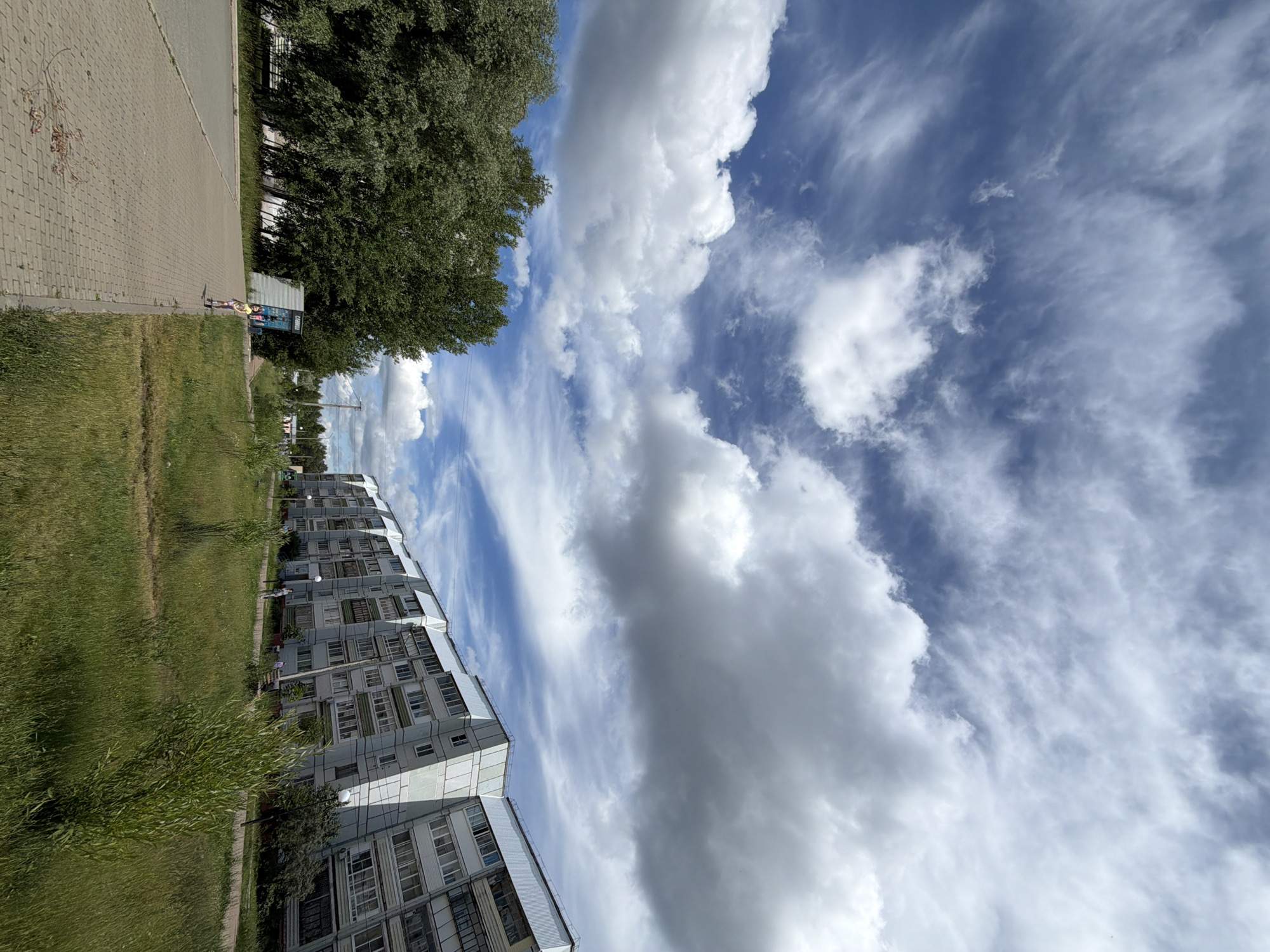 The image shows a wide, paved walkway in Chaikovsky, Russia, bordered by green grass and a line of trees on the left. A young girl in a yellow shirt and pink shorts is walking on the walkway. To the right of the walkway, there is a long, multi-story apartment building with a distinctive angular design. The sky is filled with a mix of white, puffy clouds and thinner, wispy clouds against a blue background. In the distance, more trees and buildings are visible, suggesting an urban or suburban environment.