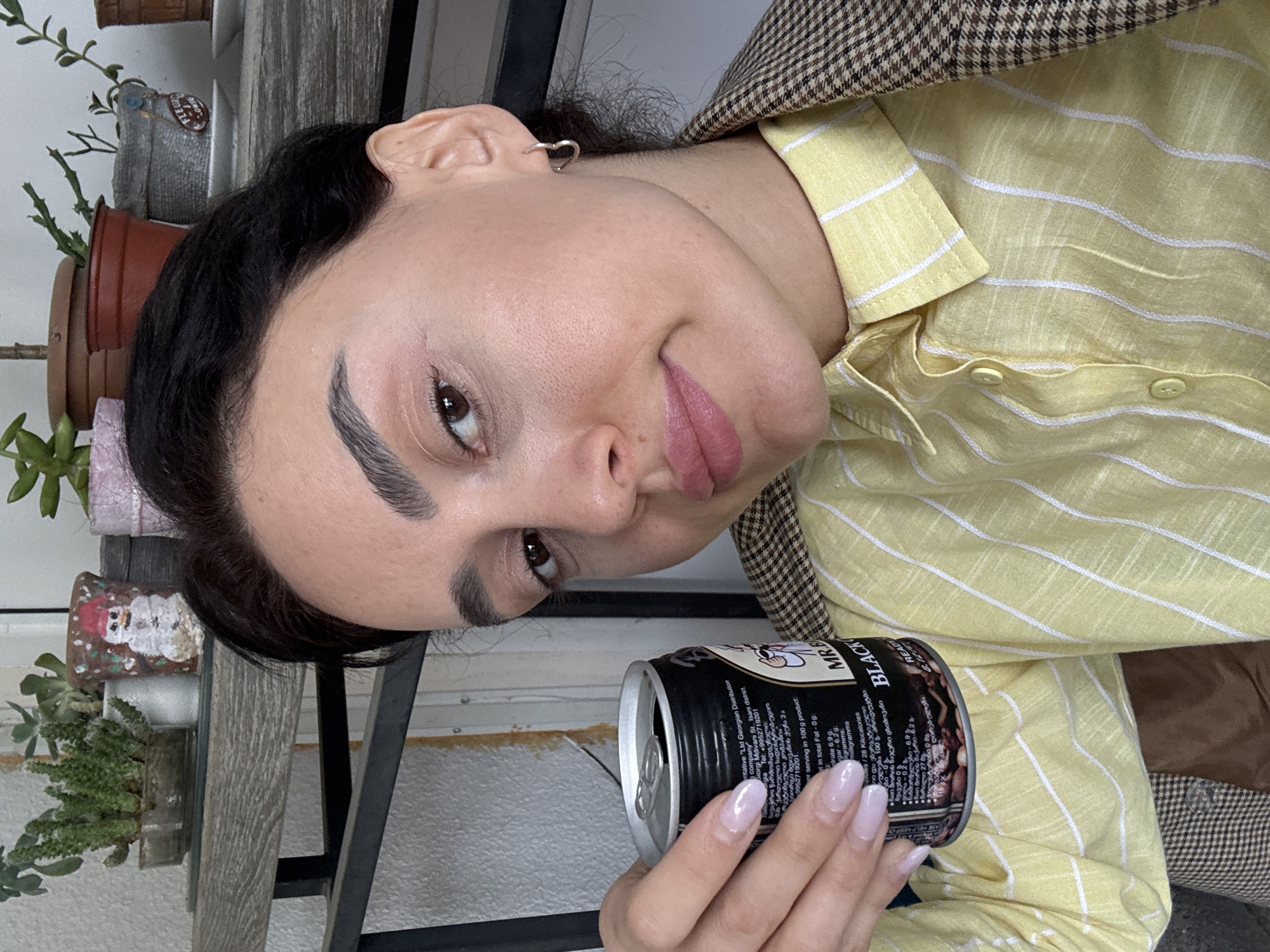 A woman with dark hair and light skin is indoors, holding a can of "MR.B Black Ready" coffee drink. She wears a light yellow, striped shirt and a brown and white plaid jacket. Her nails are painted light pink. The can's label has the Georgian Distribution company information, indicating a location in Tbilisi, Georgia. In the background, there is a shelf with potted plants and a white fence.