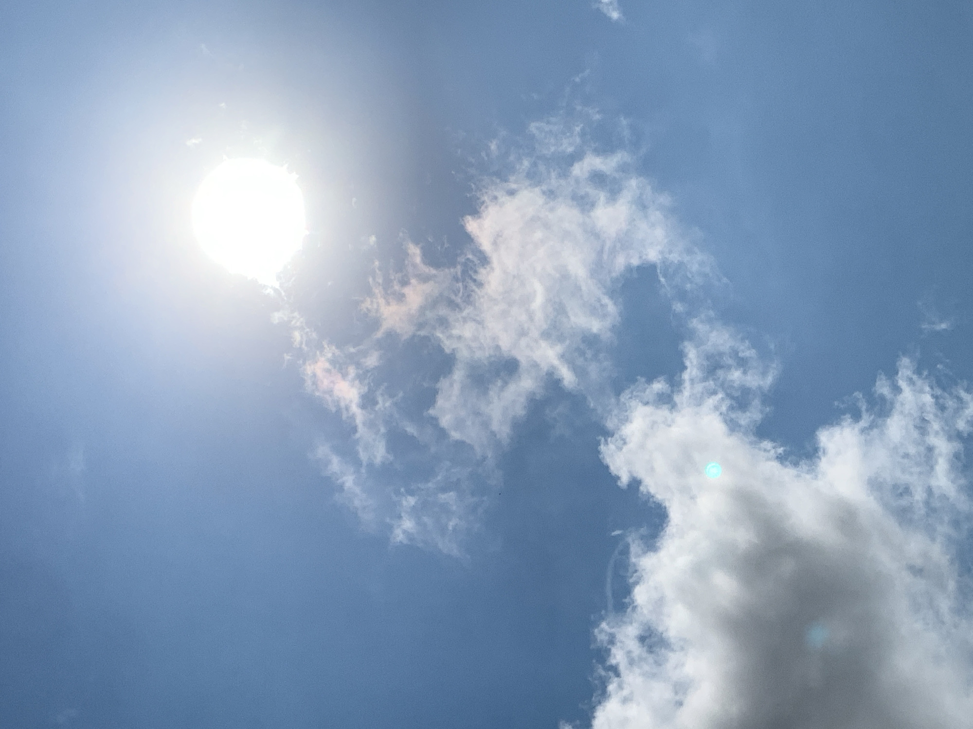 A bright sun is visible in the upper right portion of the frame, casting a strong glare. Fluffy white clouds are scattered across a clear blue sky, with some clouds showing hints of pink and orange, suggesting the time of day might be around sunrise or sunset. There is no indication of people, objects, or specific city landmarks within the frame, making it impossible to determine the exact location within Khazar Raion, Azerbaijan, or any specific actions or interactions taking place. The image is a straightforward depiction of a sunny sky with clouds.