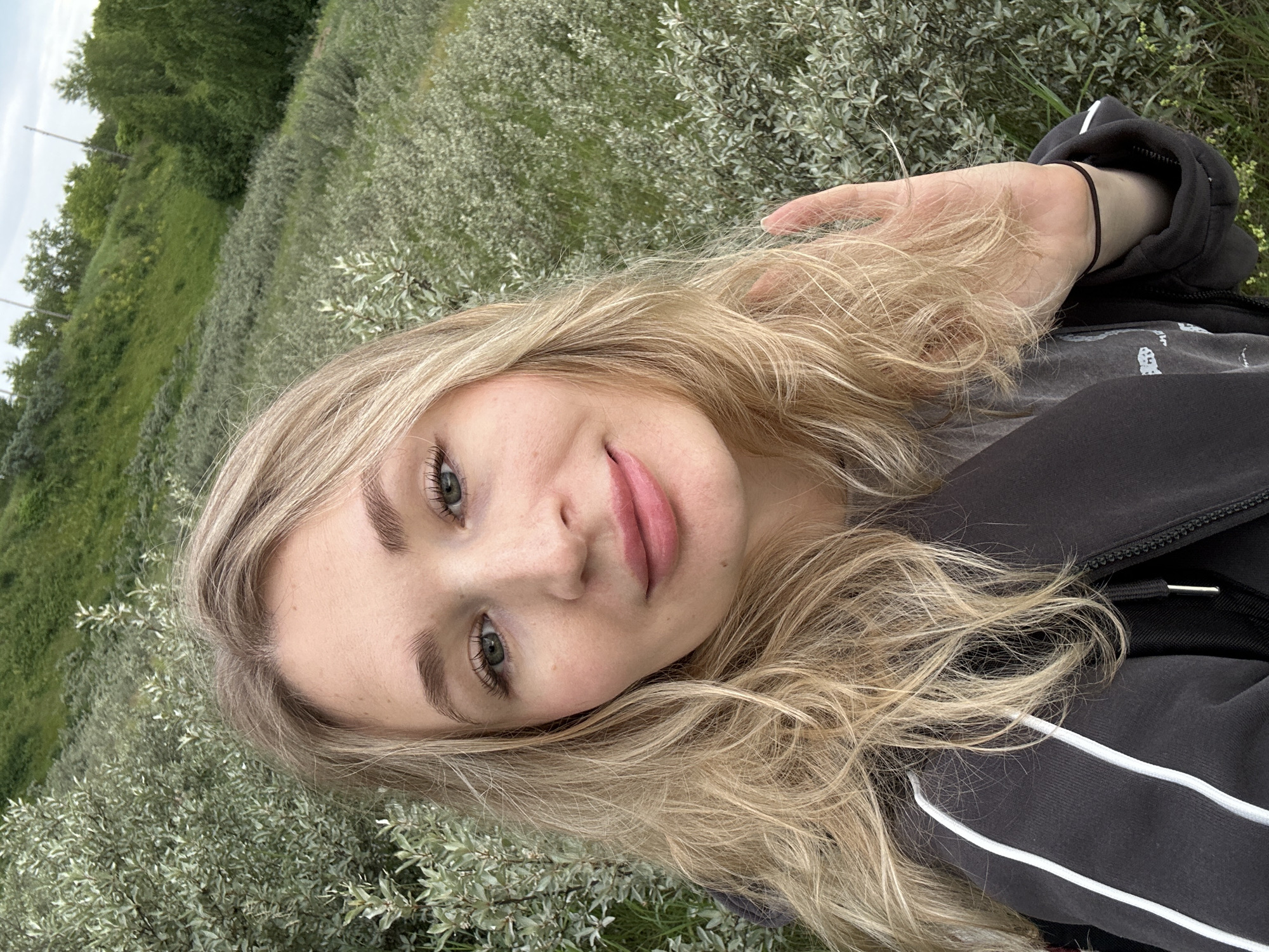 A young Caucasian woman with blonde, wavy hair is taking a selfie outdoors. She is wearing a dark grey hooded jacket with white stripes on the sleeves. The background is filled with dense, green foliage, suggesting a natural, possibly park-like or wooded area. The woman is looking directly at the camera with a slight smile. Her right hand is raised near her face, with a thin black bracelet on her wrist. The overall impression is of a casual outdoor portrait. The specific location in Chaikovsky, Russia, is a grassy hillside covered with shrubs and trees, with a visible sky in the upper right corner, indicating it's daytime.