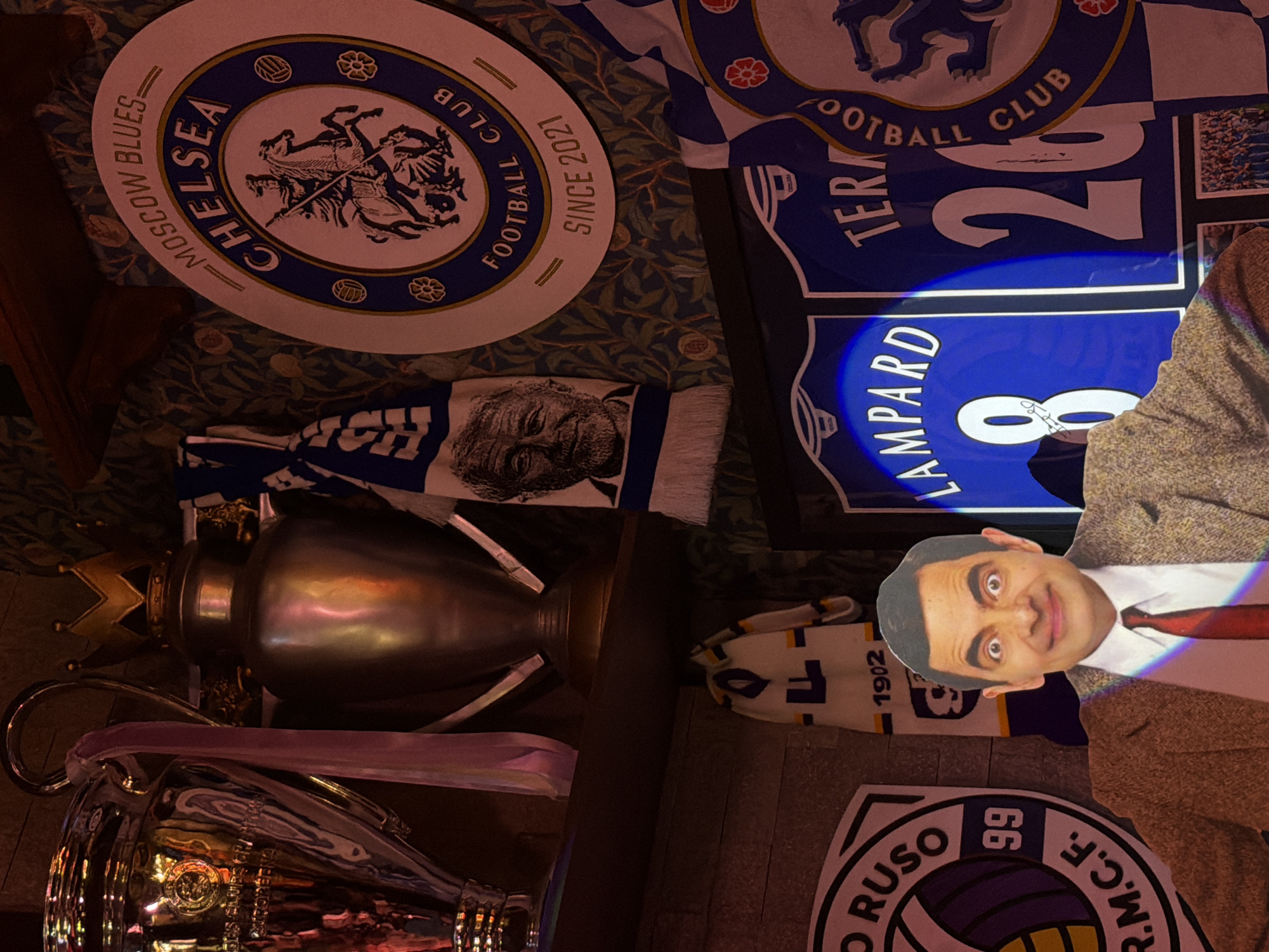 The image is a collection of Chelsea Football Club memorabilia. There's a Chelsea Football Club badge with "MOSCOW BLUES" and "SINCE 2021" printed on it, along with the words "CHELSEA" and "FOOTBALL CLUB". A scarf with a man's face on it is also visible. A replica of the Premier League trophy with a golden crown sits next to a replica of the UEFA Champions League trophy with a pink ribbon around it. The numbers "26" and "8 LAMPARD" are displayed on a blue and white banner. A white and blue scarf with the year "1902" on it is also present. A sign with the words "O RUSO" and "R.M.C.F." along with the number "99" is visible in the lower right corner. A cardboard cutout of Mr. Bean is placed in the middle of the image. Moscow, Russia is not necessarily the location; the Chelsea badge refers to Moscow as a club nickname.