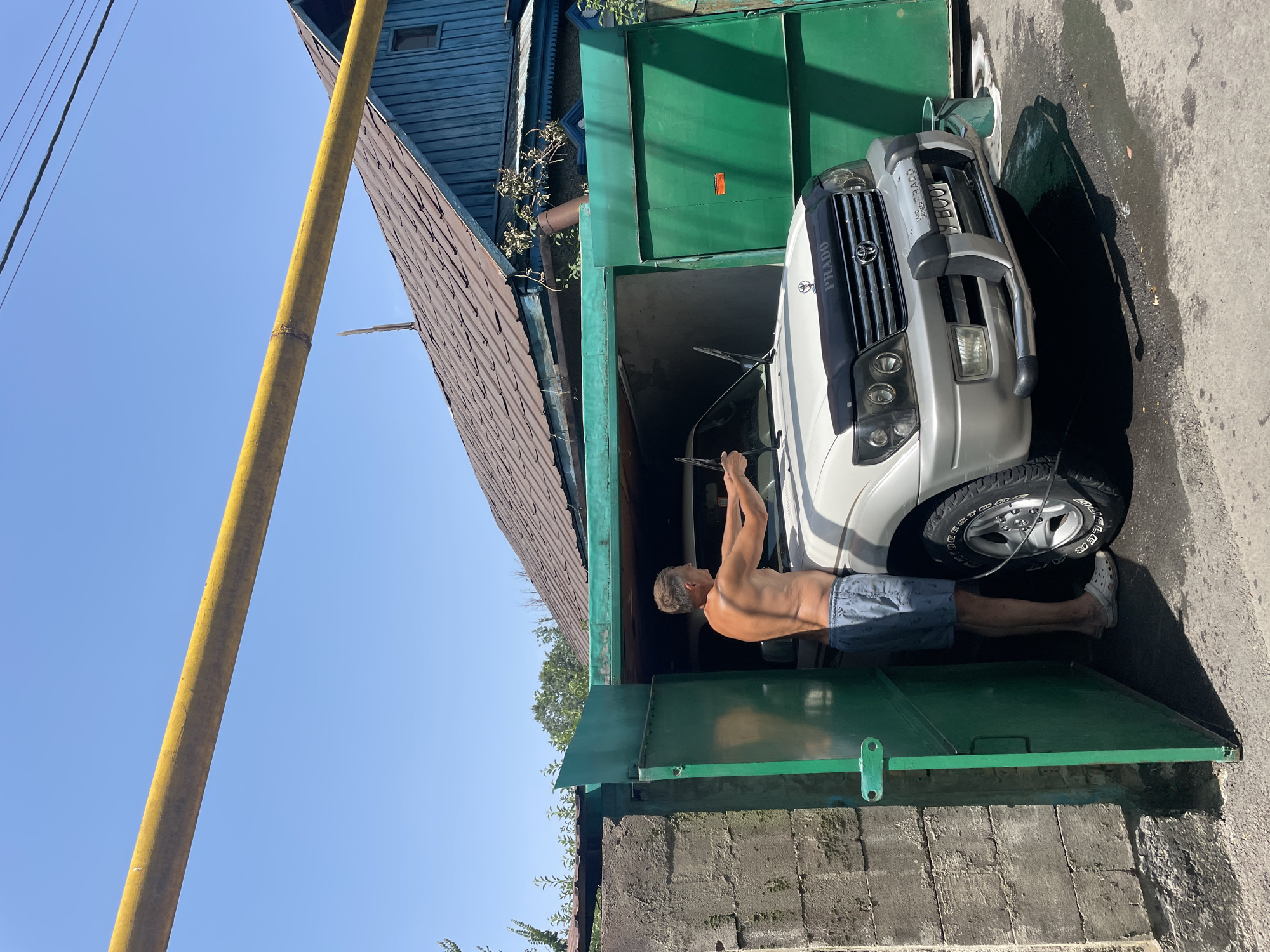 A shirtless man is washing a white Toyota Land Cruiser Prado in a garage in Almaty, Kazakhstan. The garage has green metal doors and is next to a concrete wall. The man is wearing blue shorts and white clogs. The car has a black hood deflector and a license plate that reads "7 B00". The garage is open, and the car is partially visible. There is a yellow pipe running overhead. The sky is clear and blue.