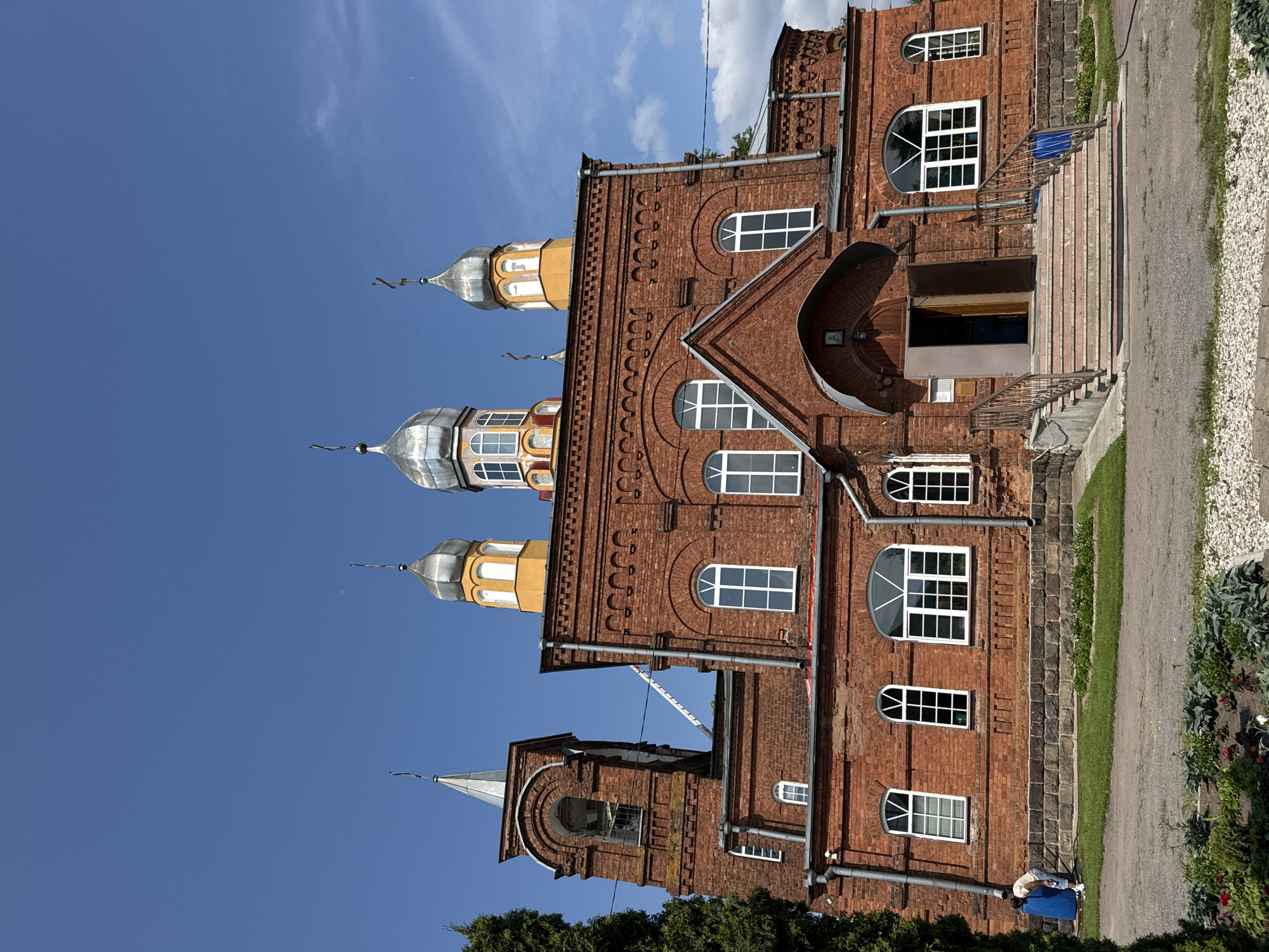 The image shows a brick Orthodox church under a clear blue sky with scattered clouds. The church has multiple onion domes, some silver and some gold and silver, topped with crosses. The facade is red brick with arched windows. A bell tower is visible on the left side of the building. There is a set of stairs leading up to the entrance, with railings. A person is visible on the left side of the building, near a large blue barrel, and appears to be bending over. Green trees and grass surround the church, with a gravel path in the foreground. The image is taken from a low angle, looking up at the church. The scene is likely the Church of the Assumption of the Blessed Virgin Mary in Chudovo, Russia, based on the architectural style and potential location.