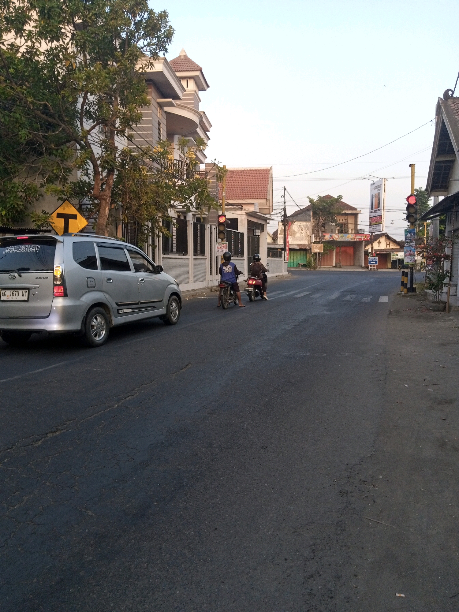 The image depicts a street scene in Cengkok, Indonesia, likely during daylight hours with soft lighting. A silver minivan is parked on the left side of the road, facing away from the viewer. The license plate is visible and reads "AG 1670 WY". A yellow diamond-shaped traffic sign with a black "T" is posted to the left of the minivan.
In the background, several buildings line the street. On the left, a modern, multi-story building with gray and white siding is prominent, partially obscured by a leafy green tree. Across the street, other buildings with tiled roofs and various signs are visible.
Two individuals on motorcycles are present in the middle of the road. The rider in the foreground is wearing a dark shirt and a helmet, and is on a red motorcycle. The rider behind them is wearing a blue shirt and a helmet, on a similar motorcycle. They appear to be stopped or moving slowly at what looks like an intersection. Traffic lights are visible, one red light is illuminated on a pole to the right of the frame, suggesting traffic control. The road surface shows signs of wear and tear, with visible cracks and patches. The overall environment suggests a typical urban or suburban street in Indonesia.