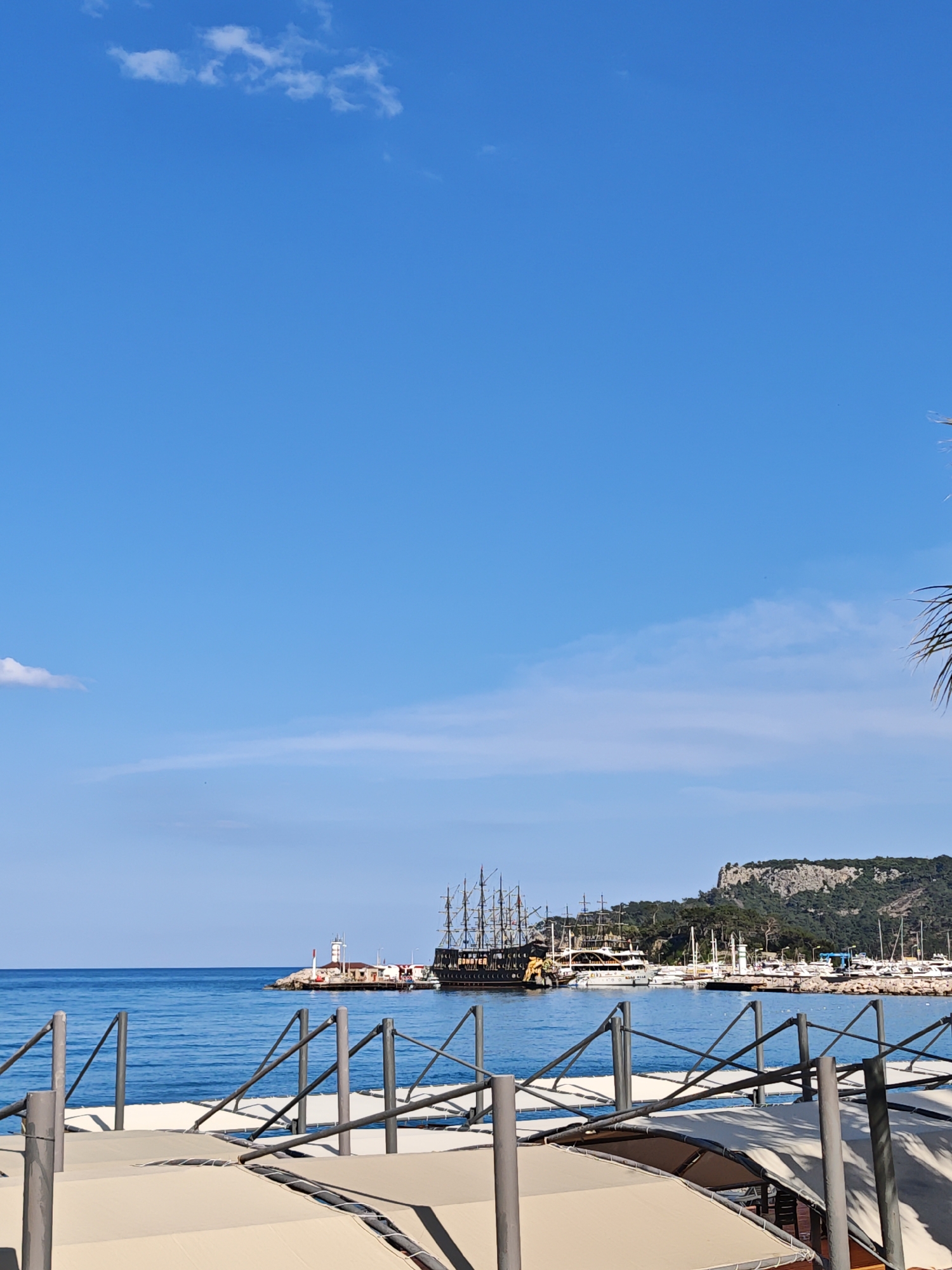 The image shows the harbor in Kemer, Turkey, on a sunny day with a clear blue sky and some light clouds. The foreground features a partially visible structure with metal railings and white awnings. In the middle ground, there is a harbor with a lighthouse, various boats, and a large pirate ship docked. In the background, there is a rocky hill covered with trees. The sea is calm and blue.
