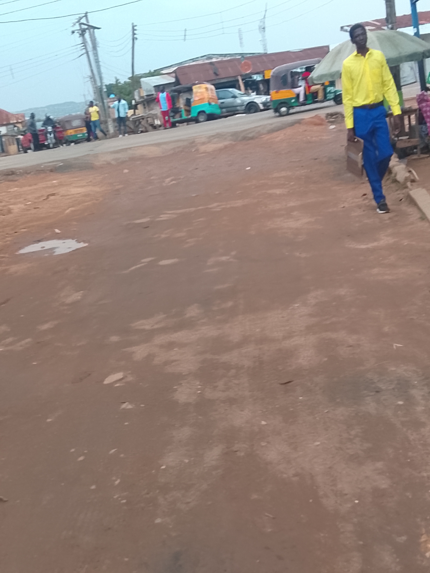 The image depicts a street scene in Jos, Nigeria. A man wearing a yellow shirt and blue pants walks to the right, carrying a brown briefcase. In the background, there are several people standing near parked auto-rickshaws ("keke napeps") and cars. The ground is dirt and reddish brown in color with puddles. Utility poles and power lines are visible. There are also simple buildings and structures with corrugated metal roofs in the background.