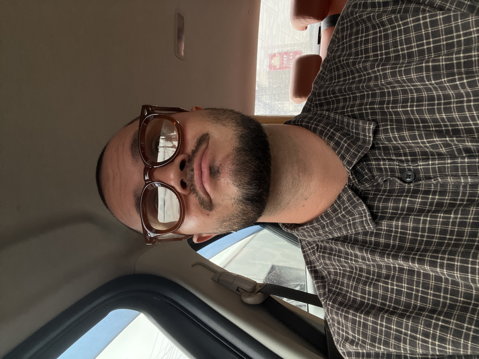 The image is a close-up, low-angle selfie of a man in a car. The man has a beard and is wearing glasses with thick, brown frames. He is looking at the camera and his mouth is slightly open. He is wearing a dark-colored plaid shirt. The interior of the car is visible, including the headliner, a seatbelt, and a window. Outside the window, a building with red signage is visible. The lighting suggests it is daytime.