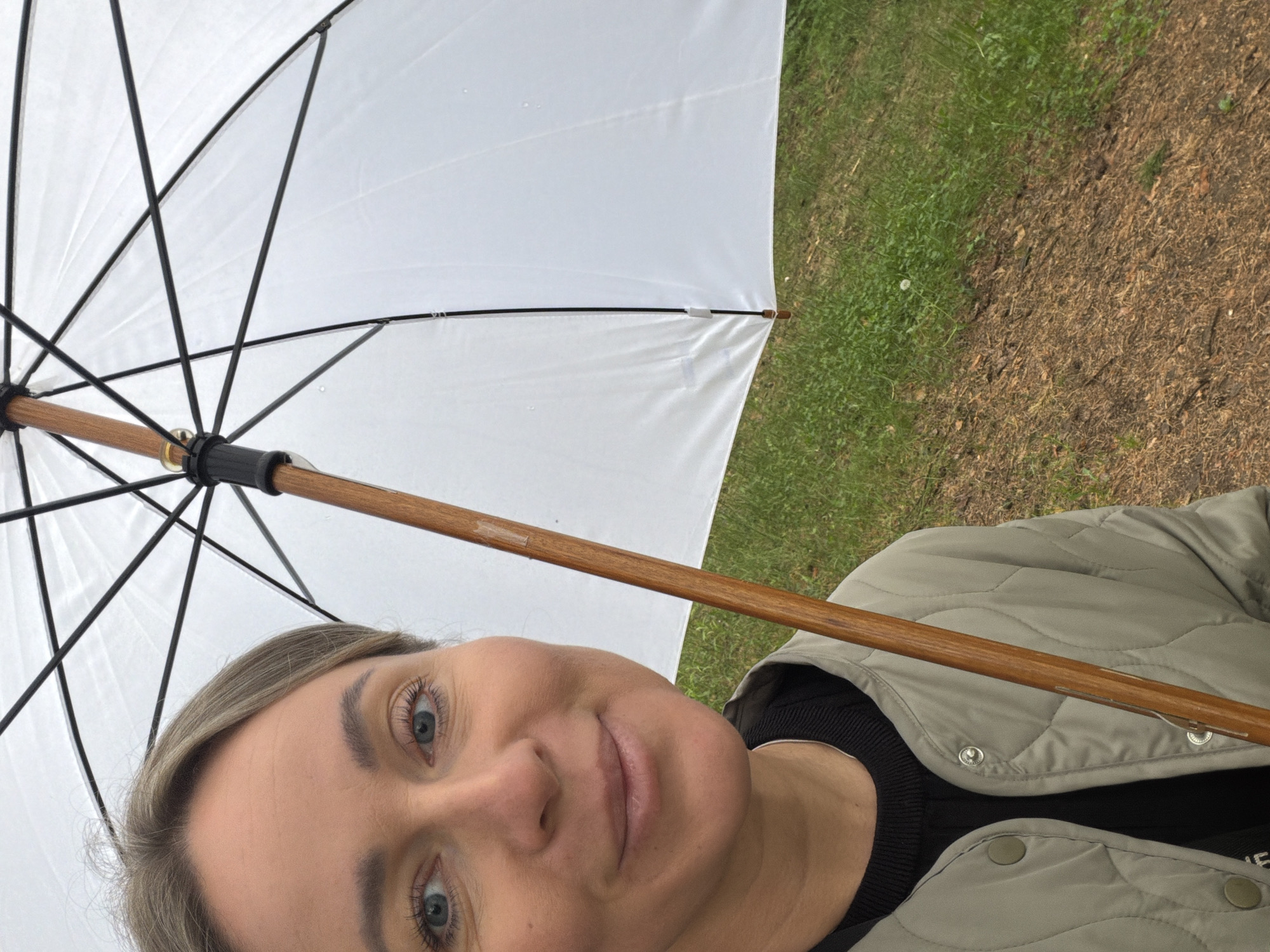 A close-up selfie captures a woman holding a white umbrella outdoors. The woman, with a fair complexion, blue eyes, and light hair, offers a neutral expression with a faint smile. She is dressed in a light green quilted jacket over a black top. The white umbrella features a wooden handle and black ribs, and small water droplets are visible on its fabric, suggesting rain or drizzle. The background of the image is divided, showcasing green grass on the left and brown earth or mulch on the right, indicating an outdoor area such as a park or path. The lighting is diffused, consistent with an overcast day. No visible text is present in the image. The location is Chaikovsky, Russia.