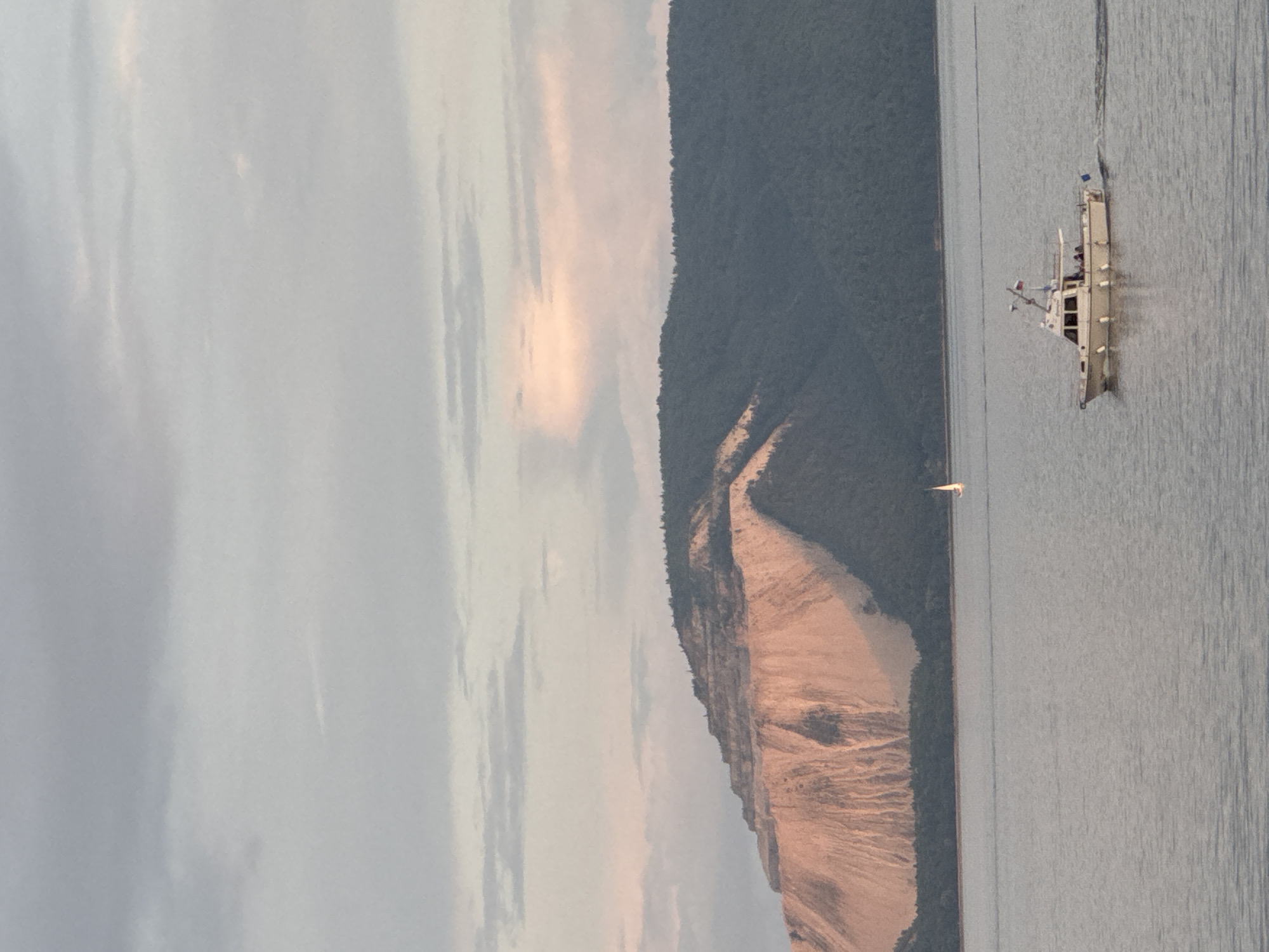 The image depicts a wide view of a body of water with a boat sailing in the foreground and a large, terraced mountain in the background. The sky is overcast with soft, diffused light, hinting at either dawn or dusk. The mountain, on the left side of the frame, features a prominent, light-colored, sandy slope that is illuminated by the setting or rising sun. The upper portion of the mountain is covered in dark green trees. A smaller sailboat is visible in the distance, to the left of the motorboat. The motorboat, on the right, is white with a red flag and appears to be carrying a few passengers. The water is calm and reflects the muted colors of the sky and the mountain. The scene suggests a serene natural landscape.